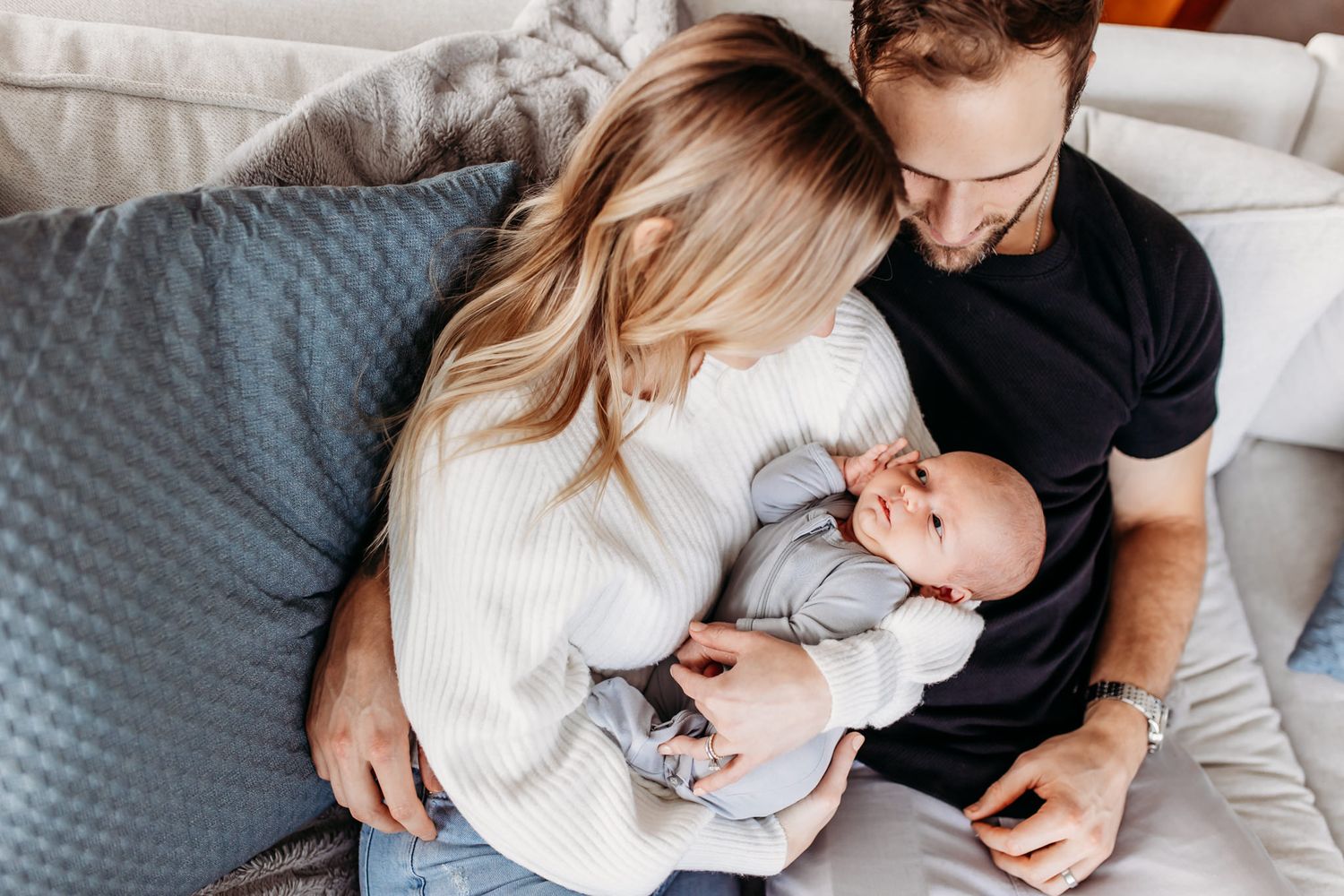 Parents sitting on gray couch while holding newborn baby in intimate lifestyle portrait.