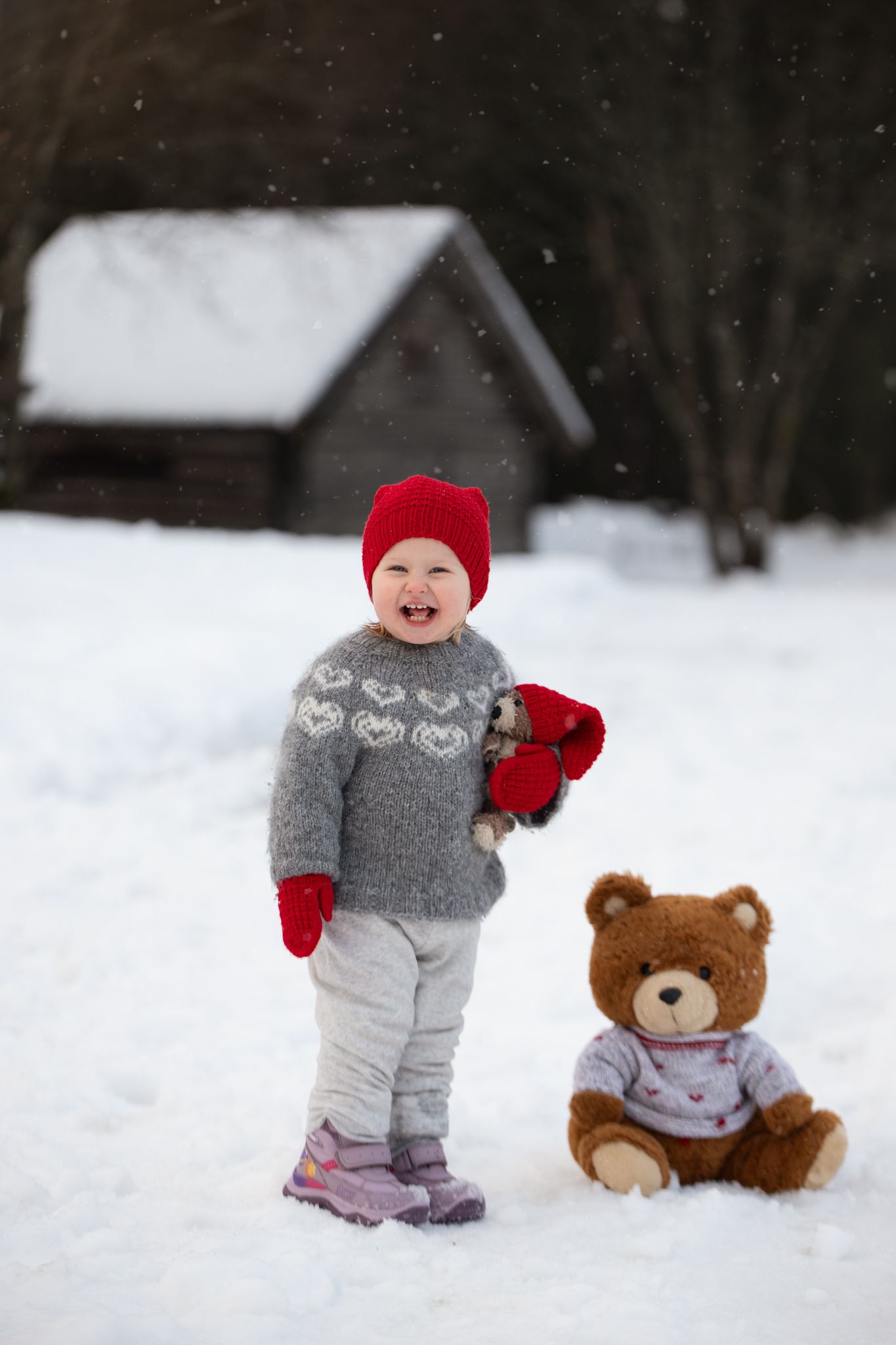 A small child in a gray outfit and red hat stands next to a teddy bear in snowy winter scenery.
