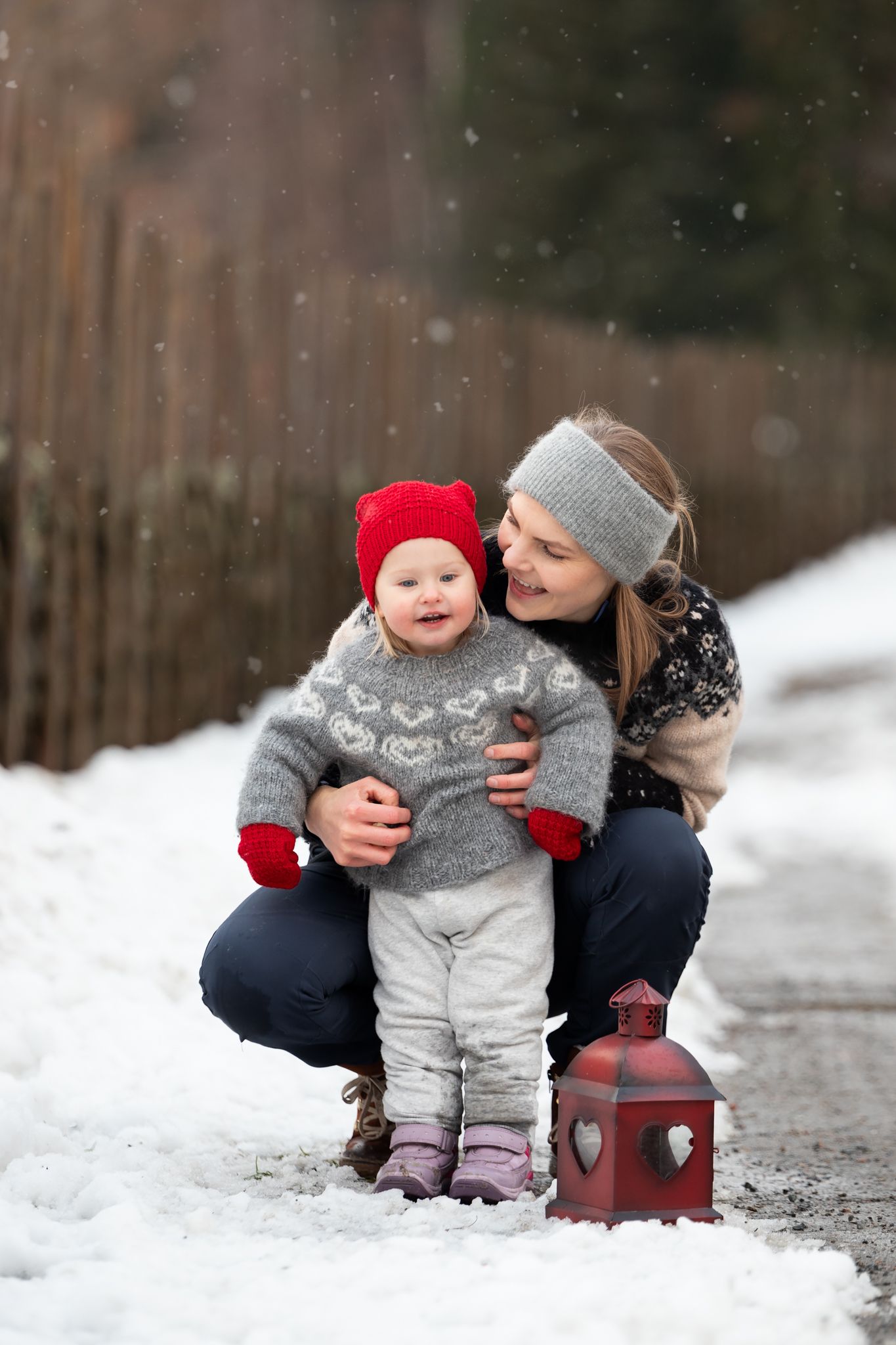 A child in winter clothing sits in the snow while being hugged from behind.
