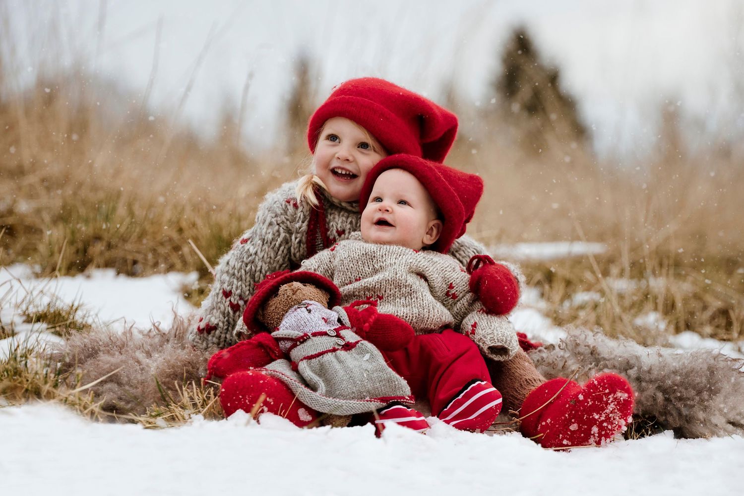 Small figure in red winter attire lying playfully in snowy field with dry grass.