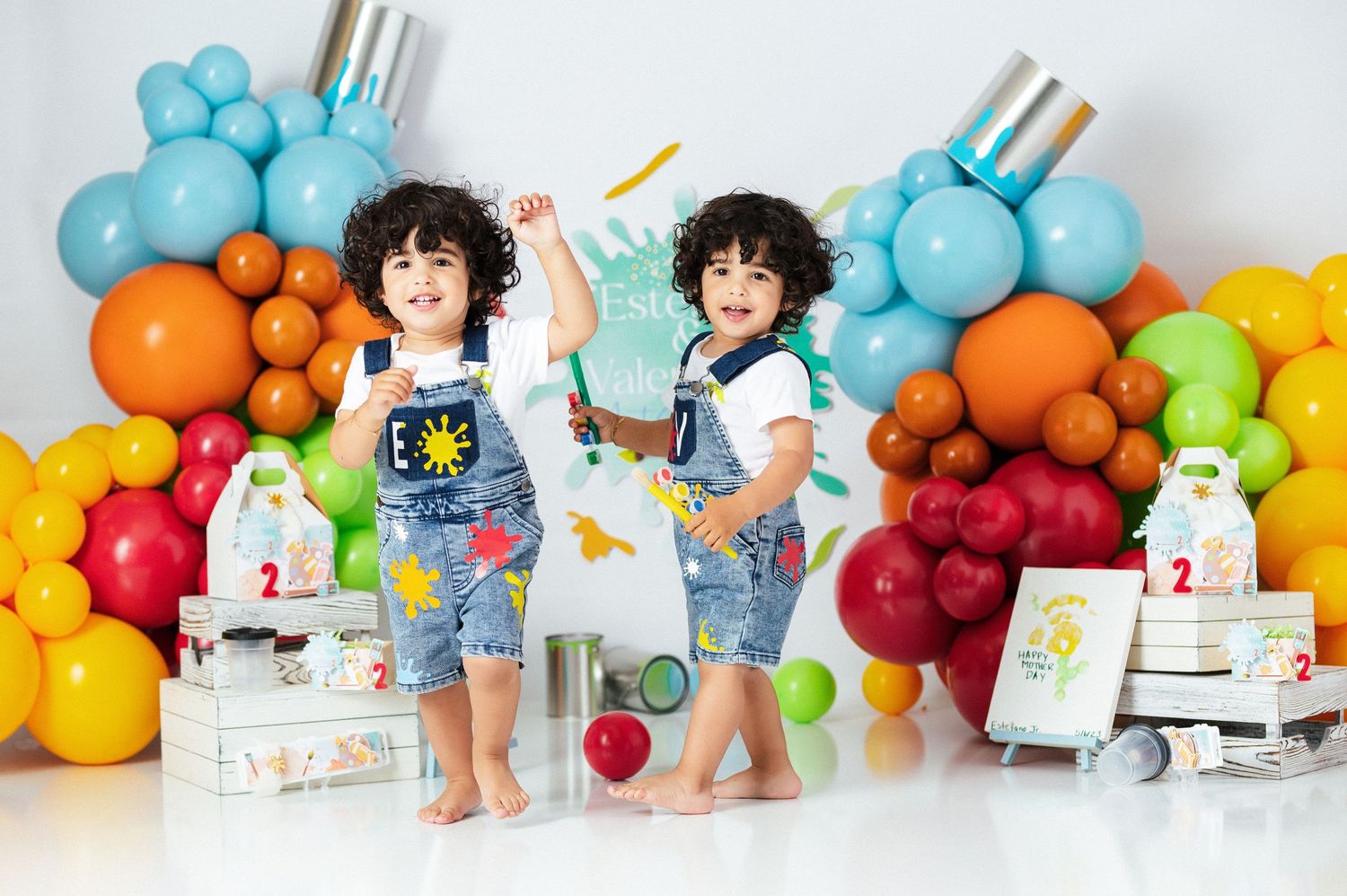 Two young twins in denim overalls posing at colorful art-themed birthday party.