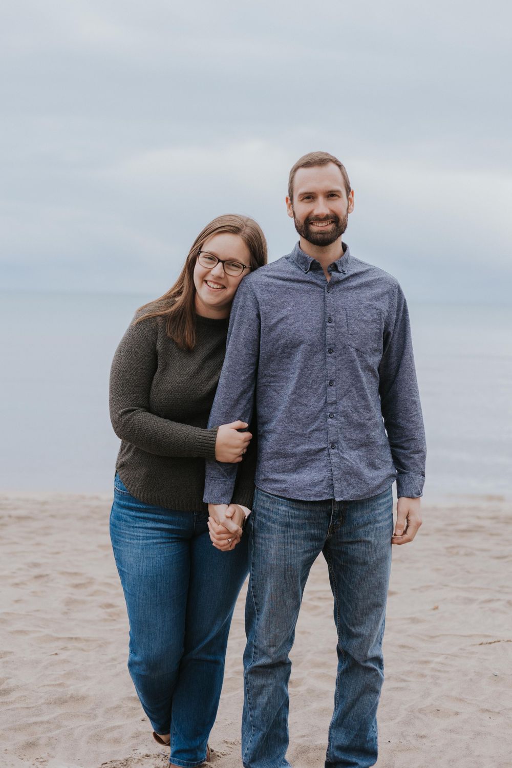 A couple poses together on a beach with overcast skies in the background.