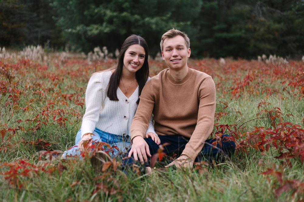 A couple sits together in a field of red autumn flowers with pine trees in the background.