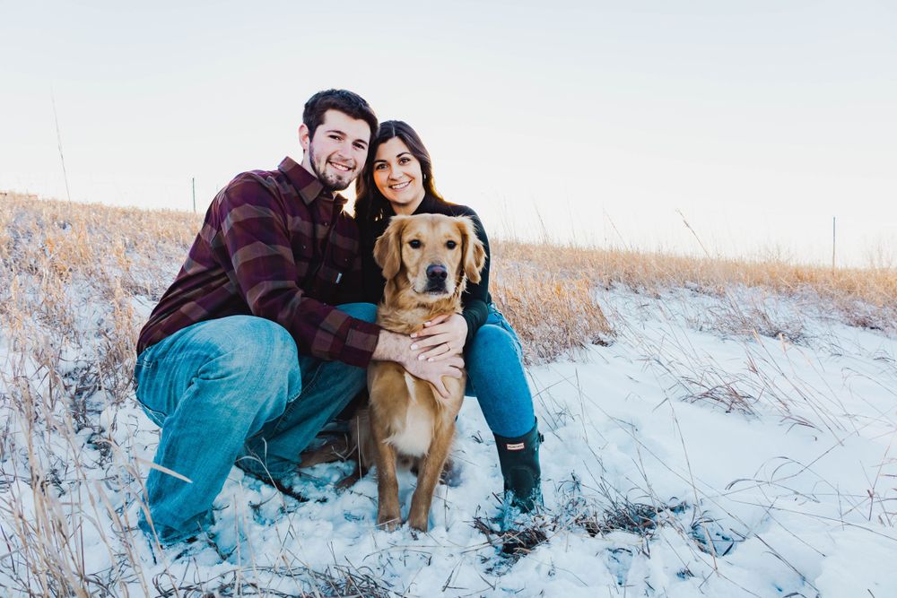 A couple sits with their golden retriever on snowy ground during a winter photoshoot.