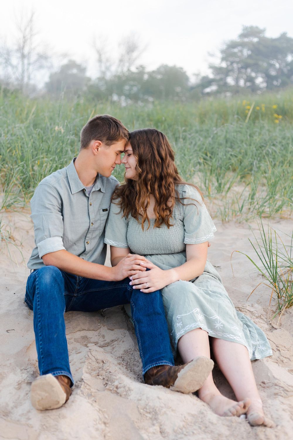 A romantic couple sits together on sandy dunes surrounded by beach grass.