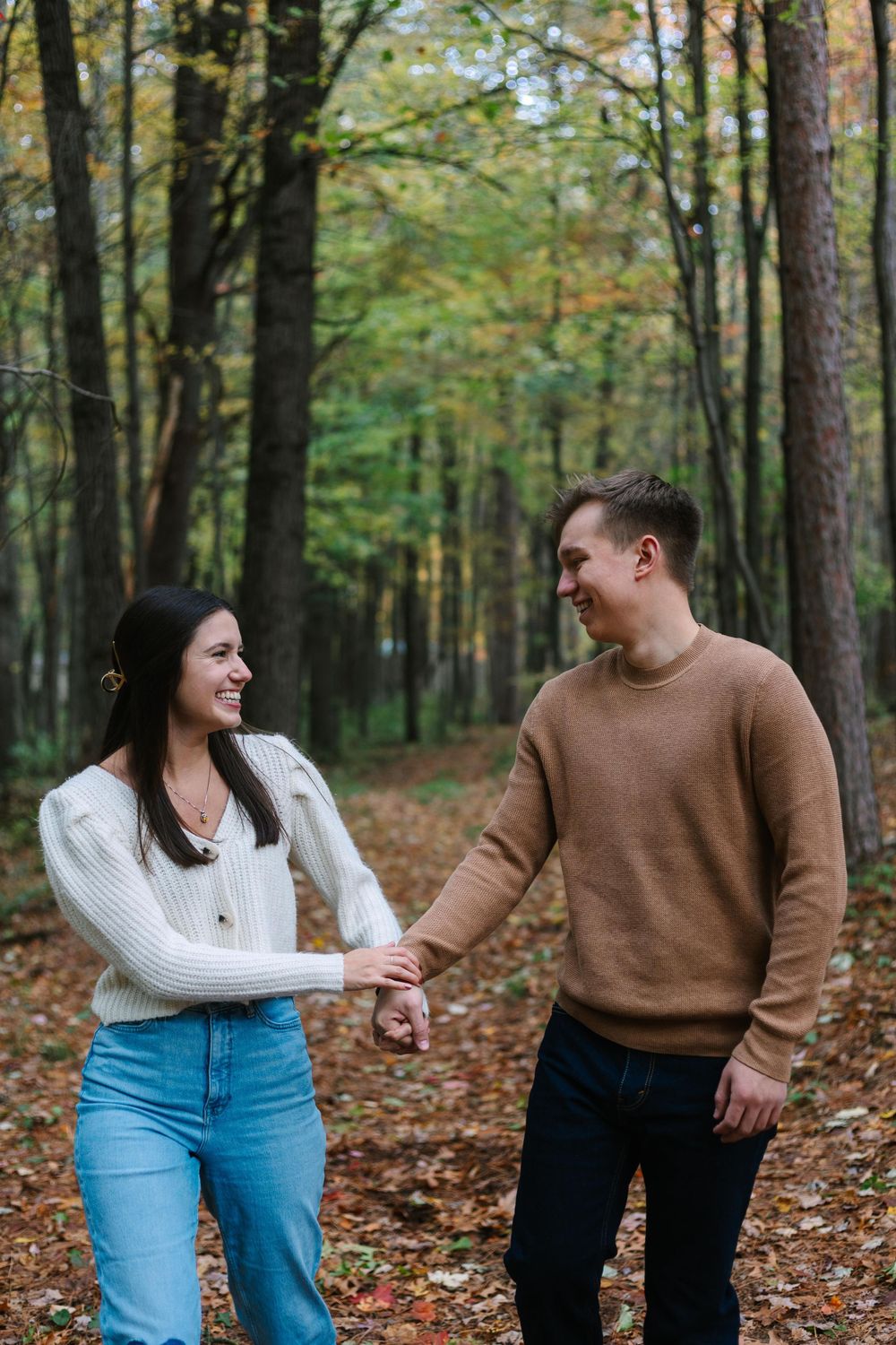 A couple holding hands and laughing together on a leaf-covered forest path in autumn.