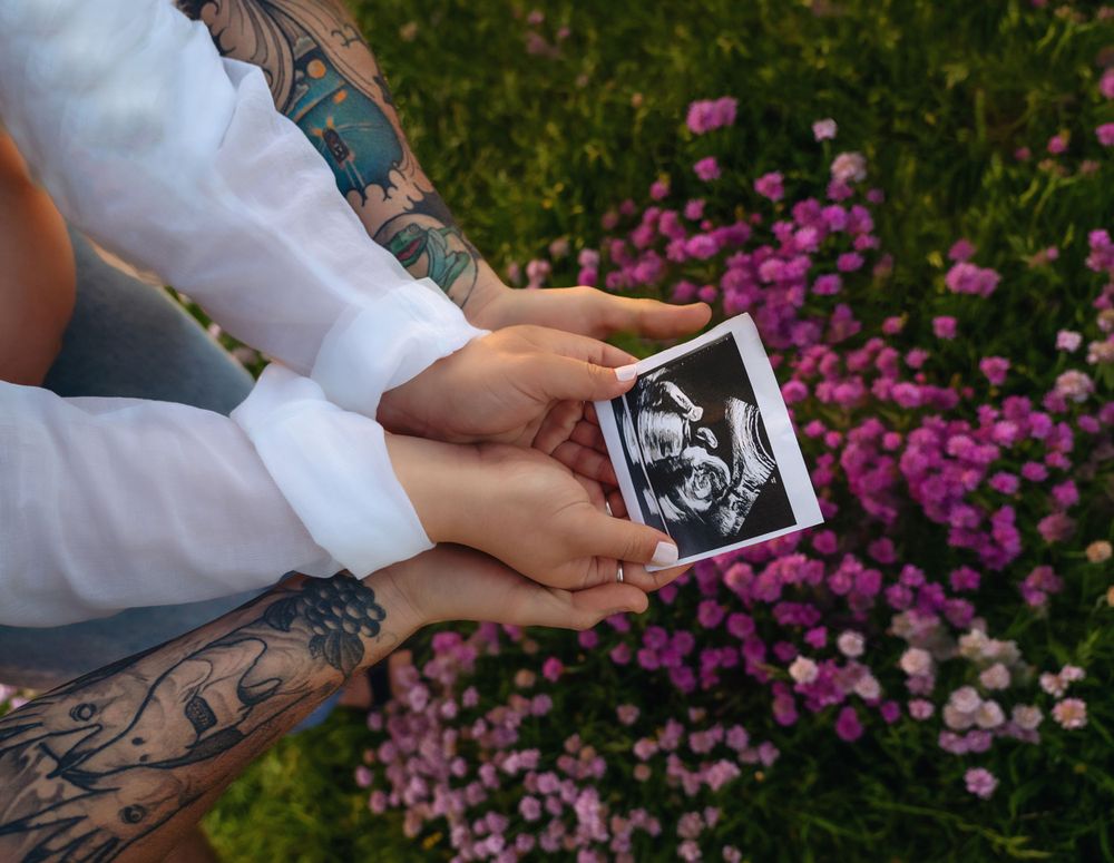 Parents' hands holding the sonogram photo, in the background there are pink flowers. Location: Milford on Sea, Hampshire.