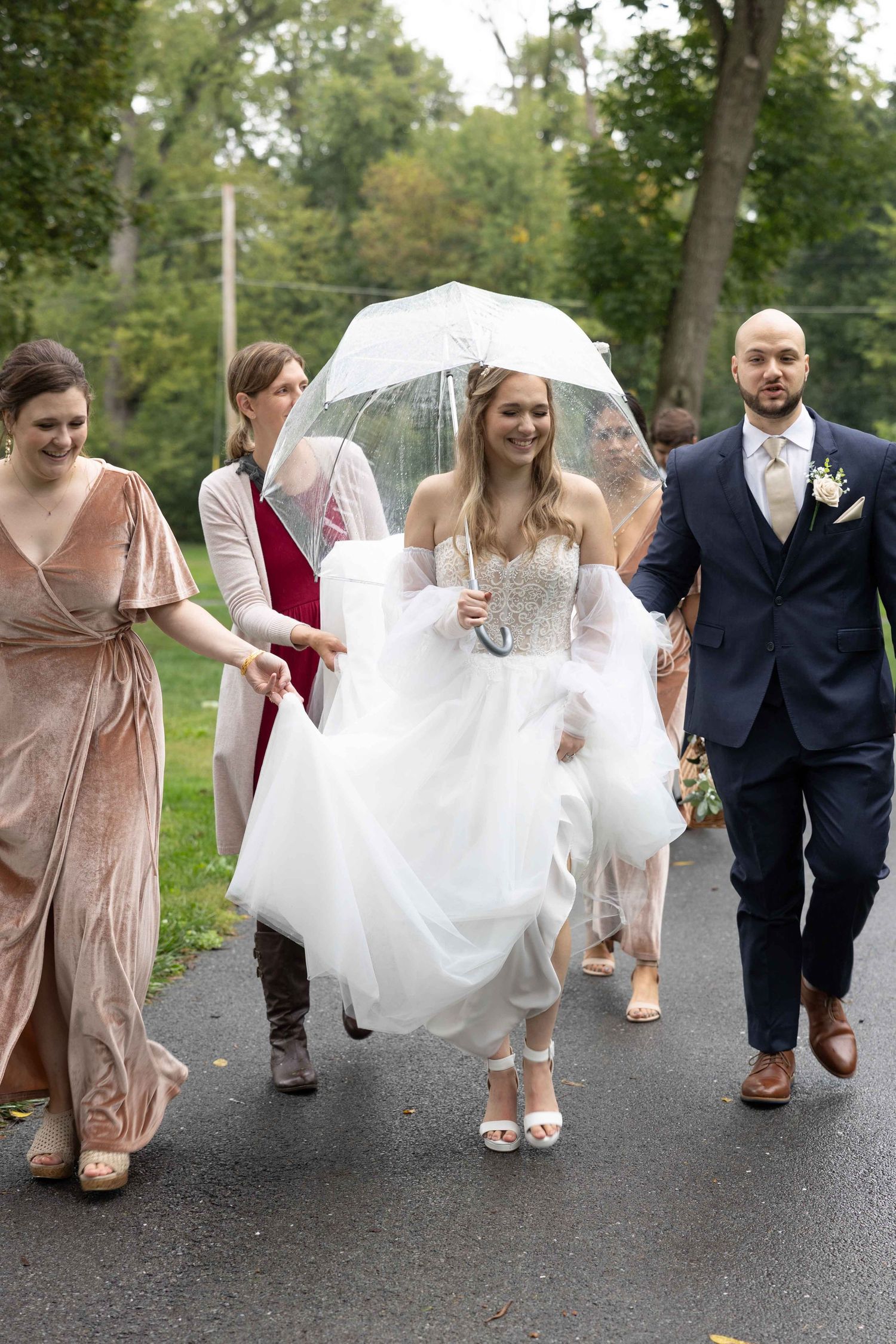 A wedding party walks down a path with the bride in white holding a clear umbrella while accompanied by attendants.