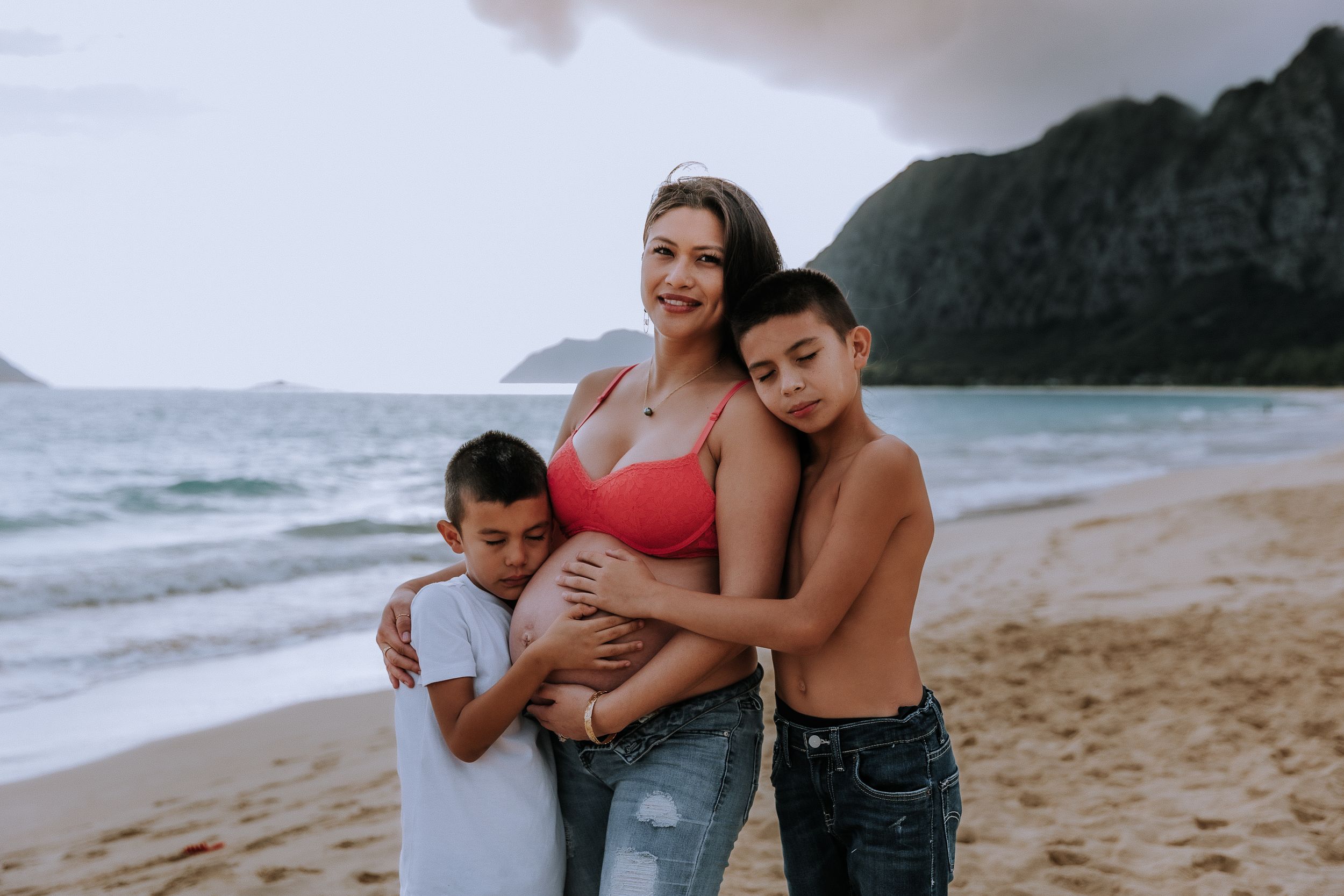 Family embraces on tropical beach with mountains and ocean in background.
