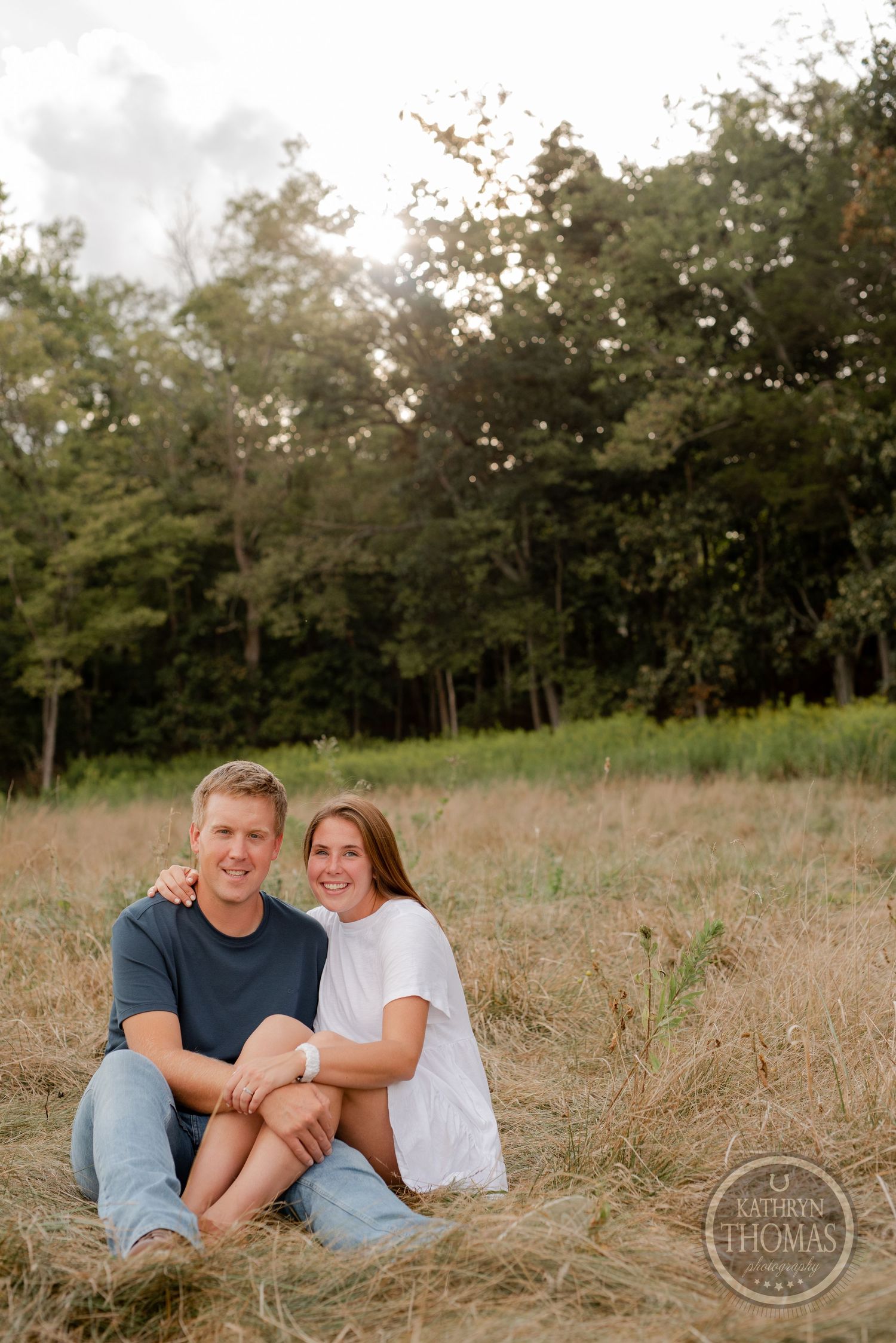 A couple sits together in tall grass with a wooded backdrop during sunset.