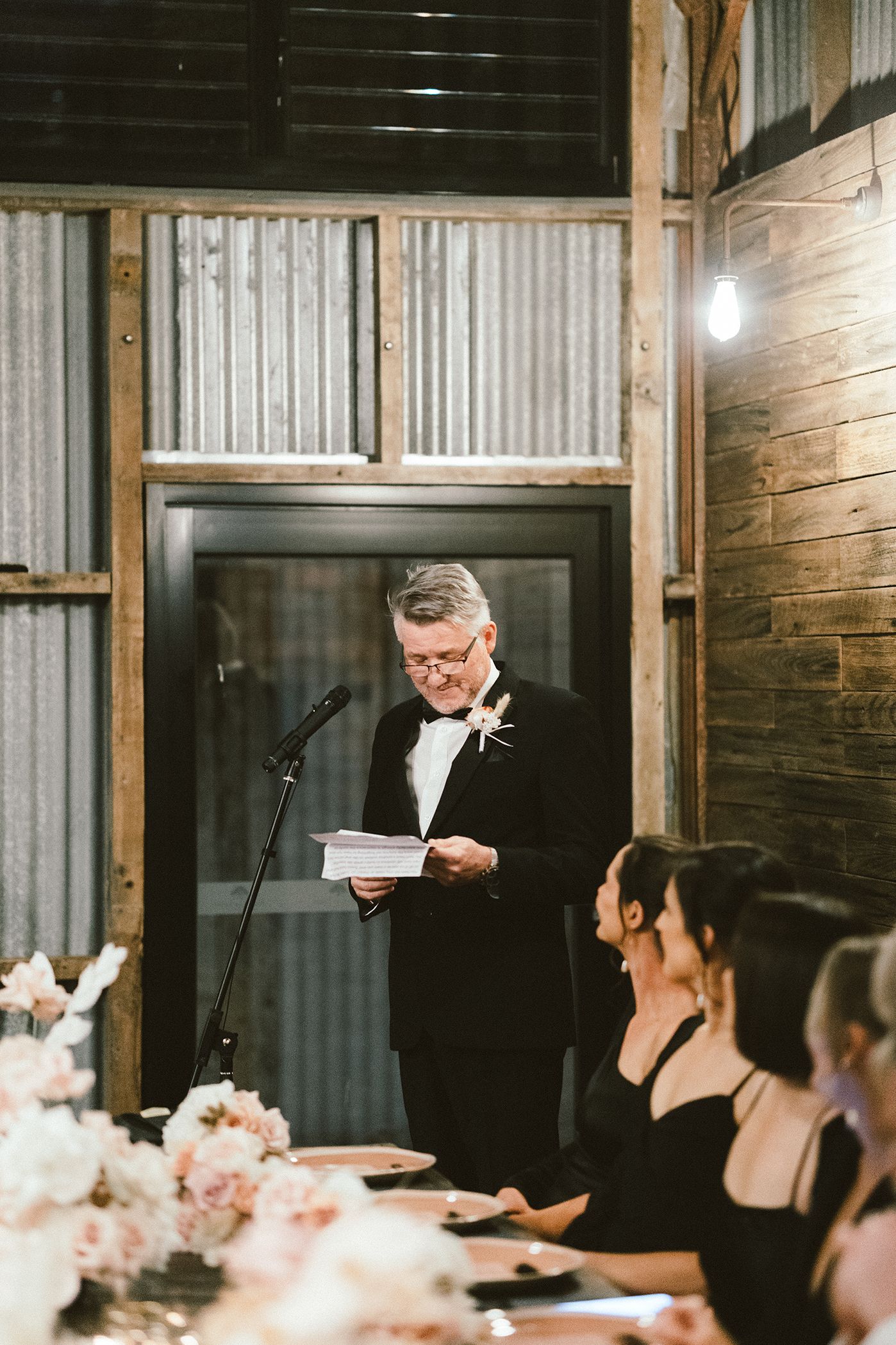 Wedding speech being delivered at rustic indoor reception with exposed brick walls and floral centerpieces.