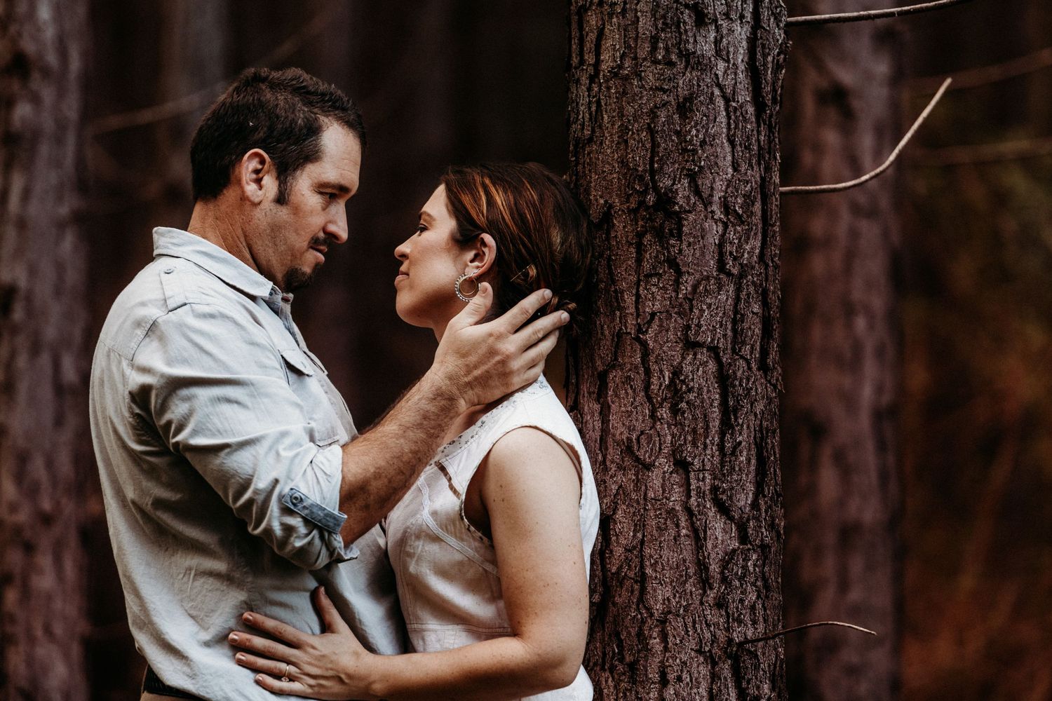 A couple shares an intimate moment against a tree trunk in a moody forest setting.