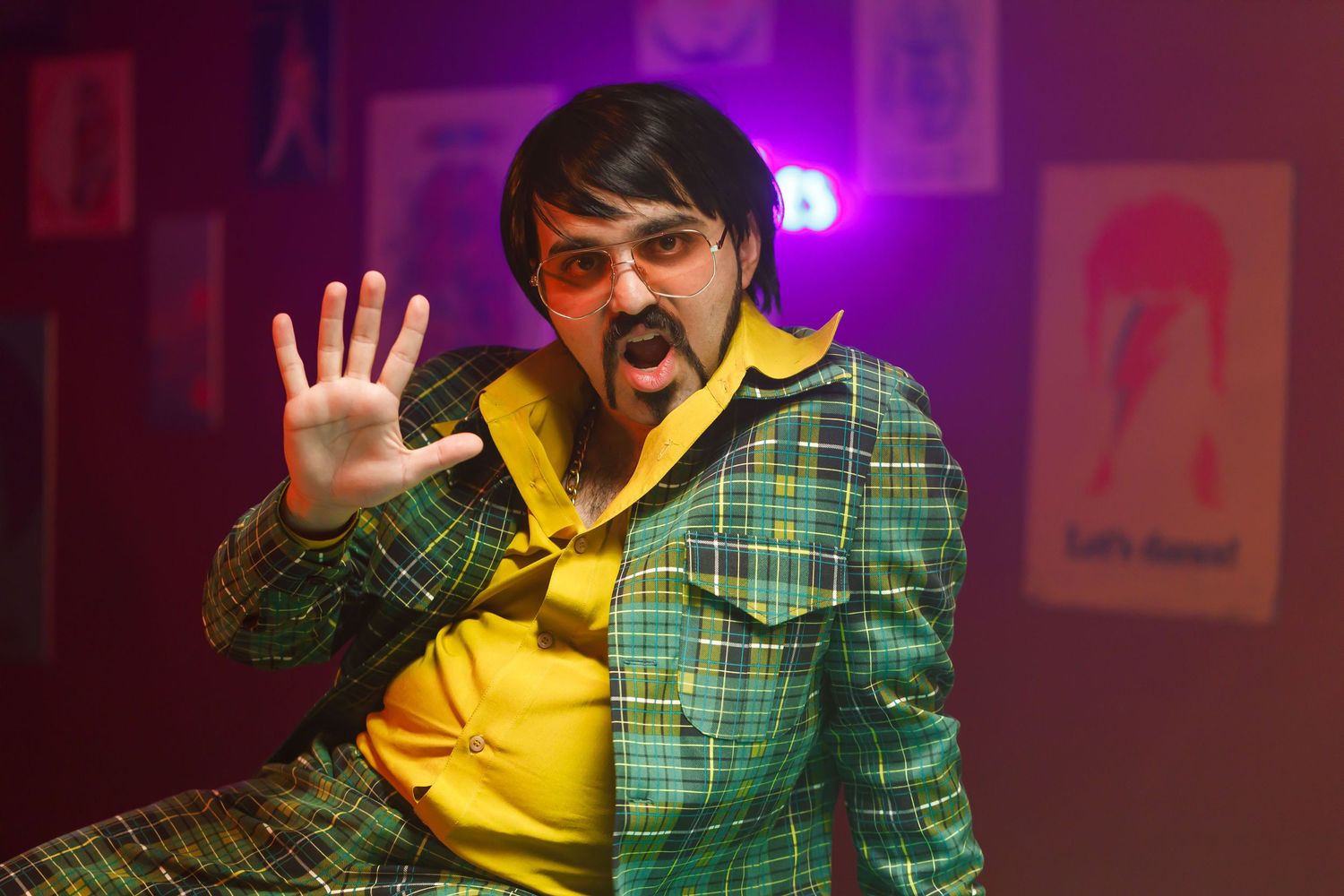 Person in yellow hoodie and plaid jacket making animated gesture under purple lighting.