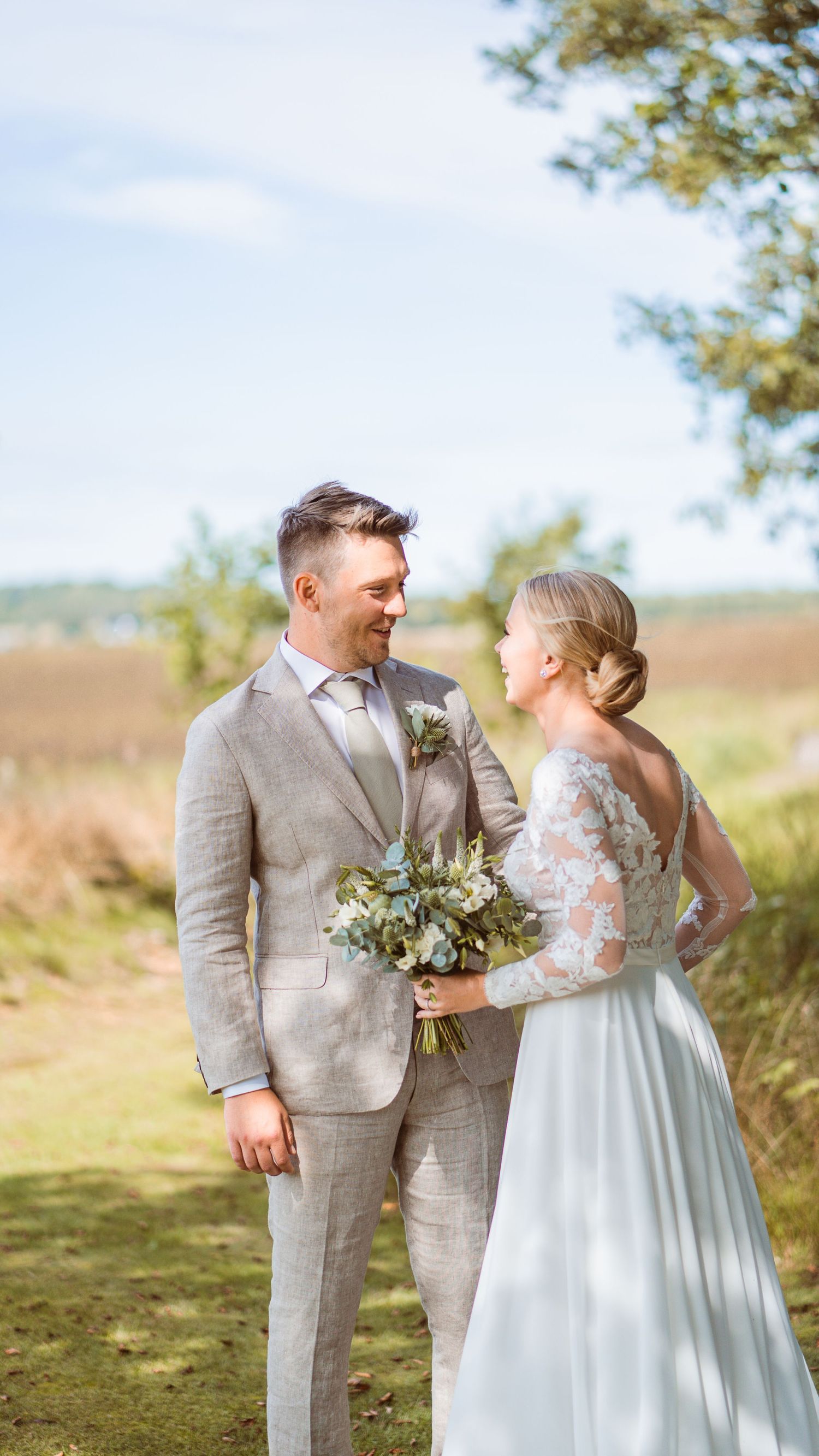 Bride and groom share a joyful glance outdoors, she in lace gown, he in beige suit, holding bouquet.