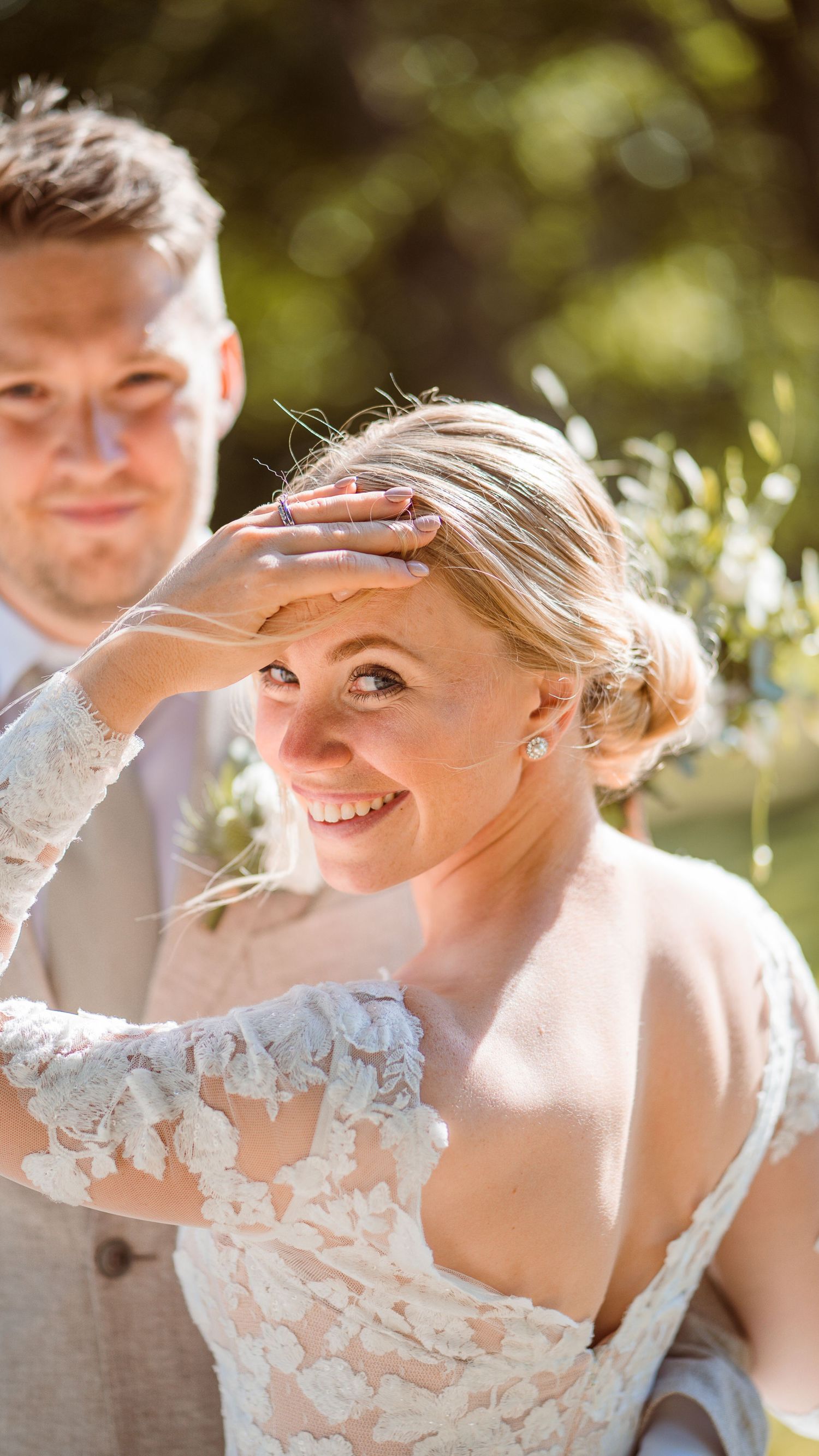 Smiling bride in lace wedding dress shielding her eyes from sunlight with groom in beige suit beside her.