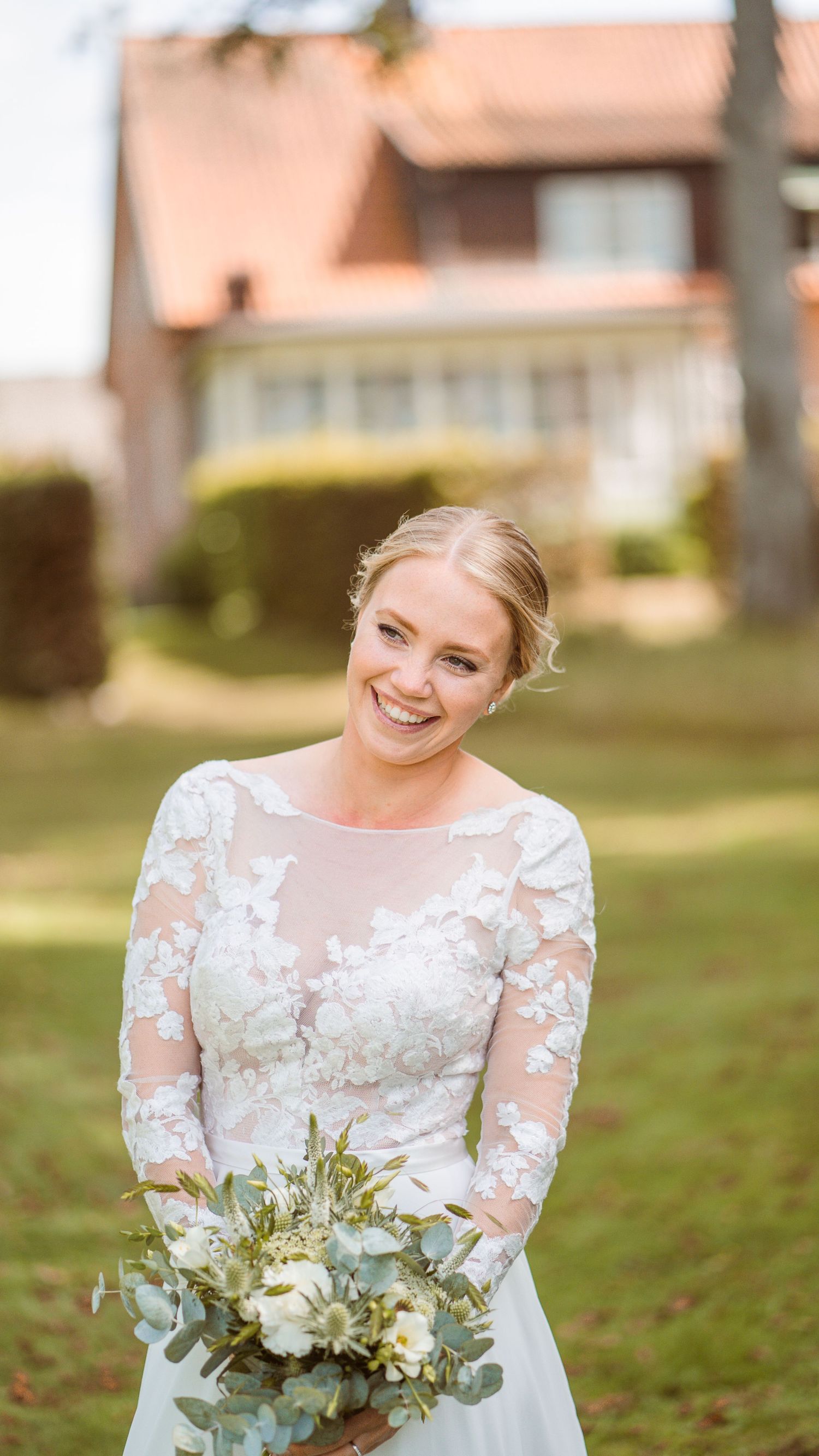 Radiant bride in long-sleeve floral lace gown smiles holding white and eucalyptus bouquet outside brick manor.