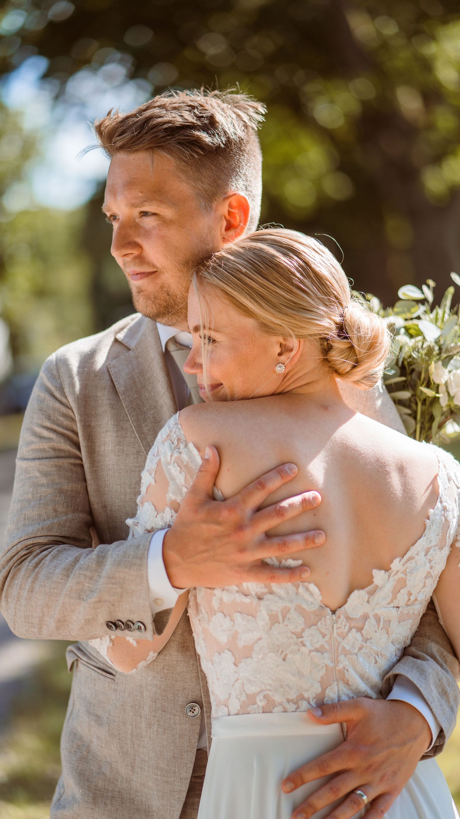 Groom in grey suit tenderly embracing bride in lace wedding dress from behind outdoors.