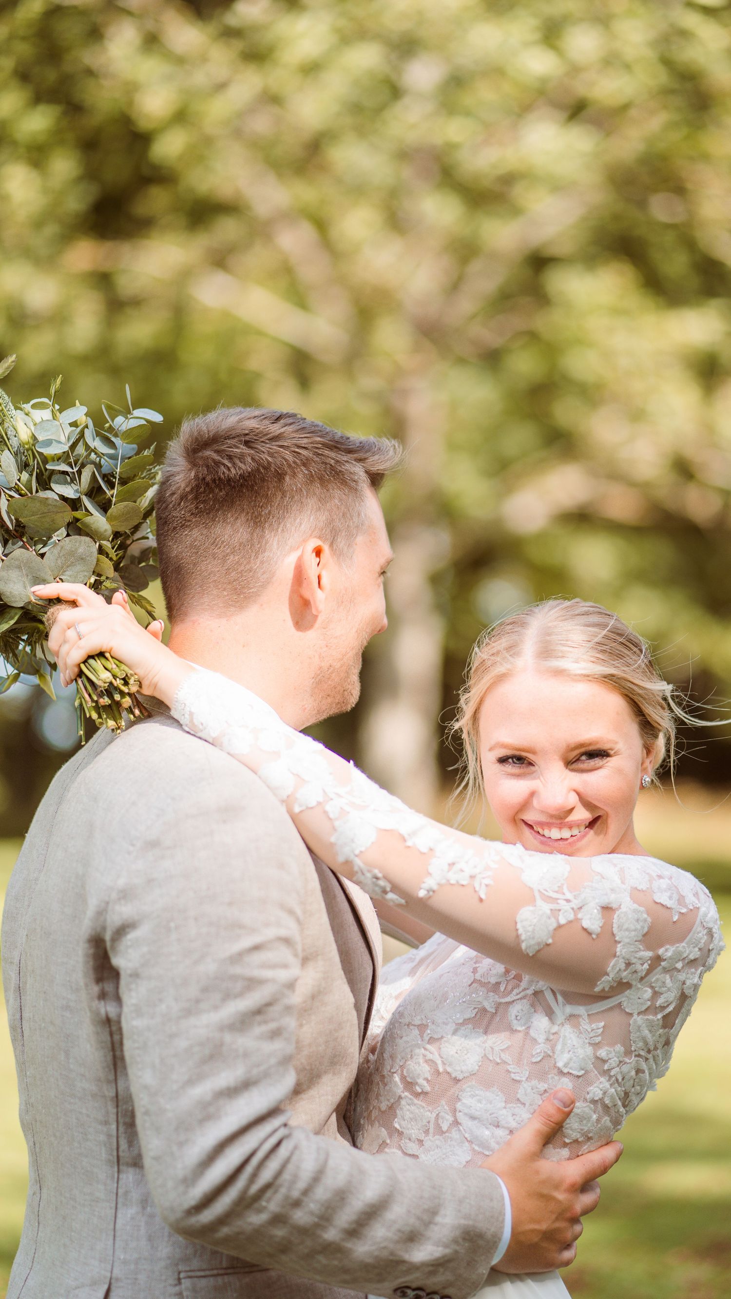Bride in long-sleeve lace gown smiles over shoulder while groom holds greenery bouquet in sunlit garden.