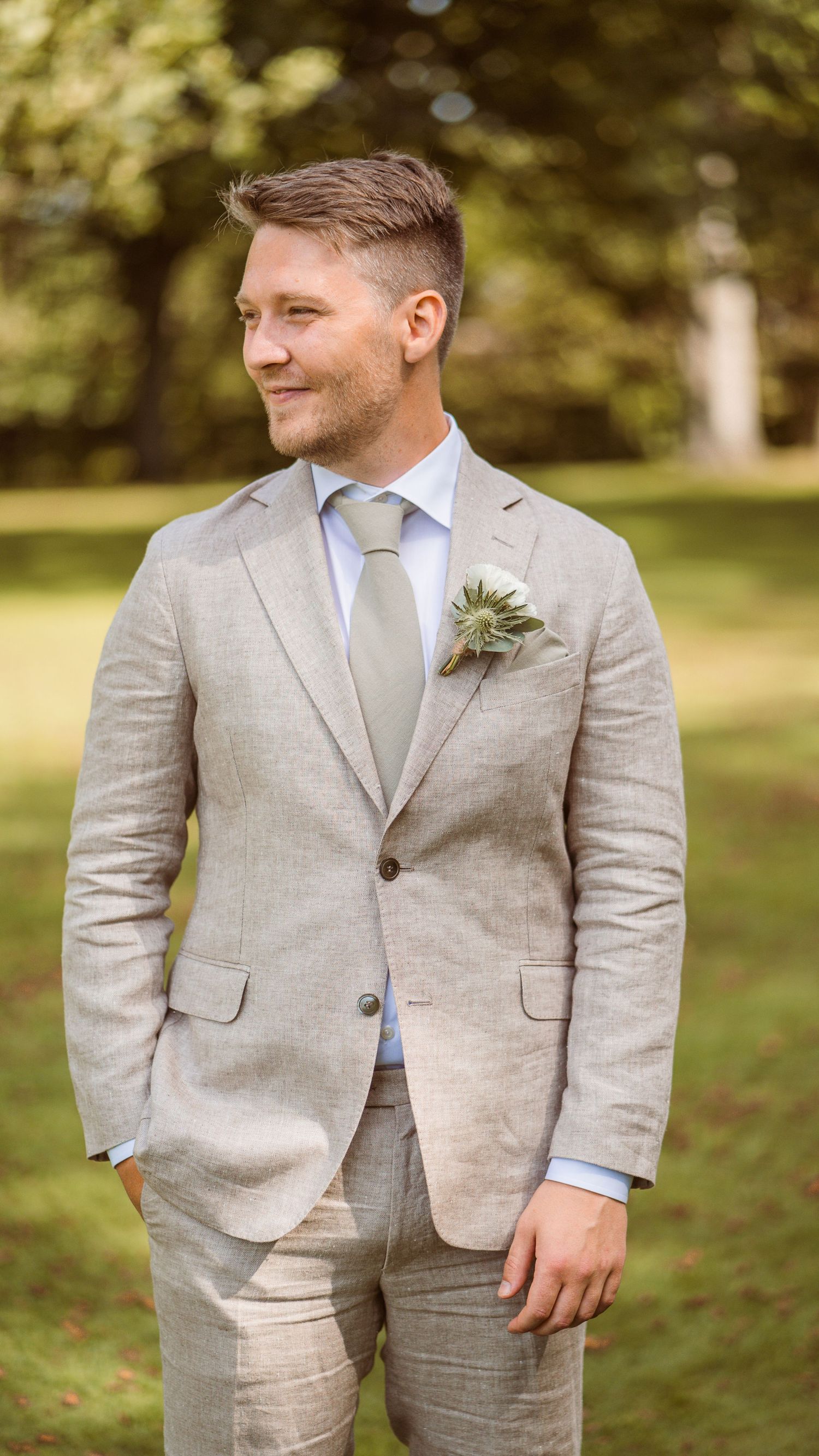 Handsome groom in tailored beige linen suit with thistle boutonniere stands confidently in lush green garden.