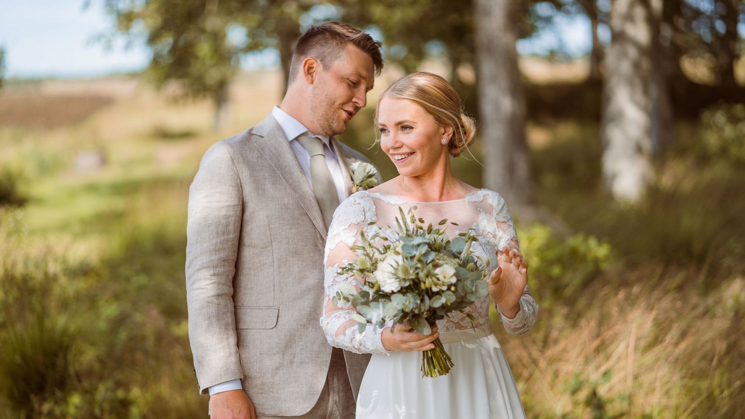 Couple in wedding attire smile warmly together outdoors, bride holding white and greenery bouquet in soft light.