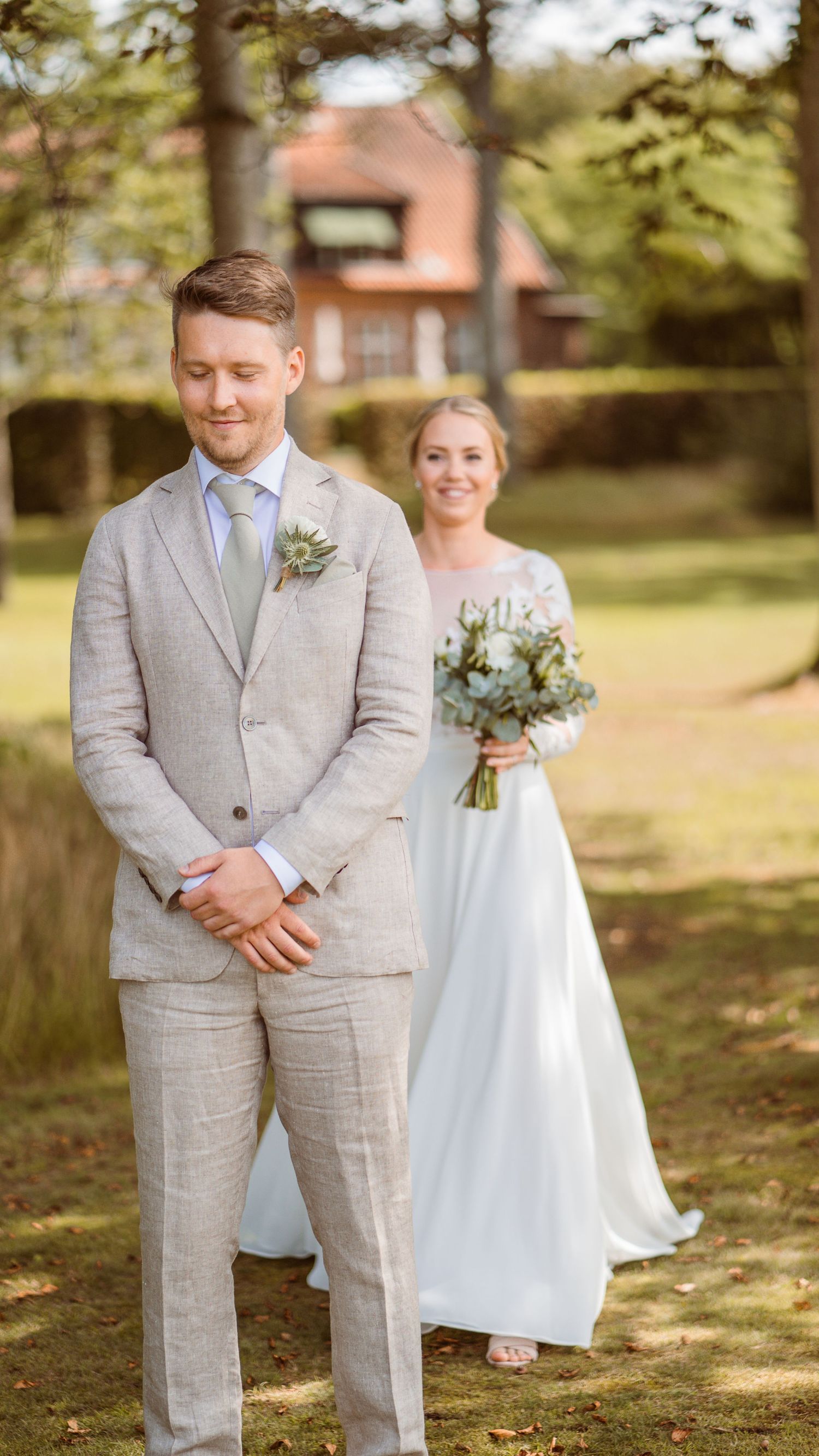 Smiling bride holding greenery bouquet sneaks up behind groom in tan suit during first look wedding moment.