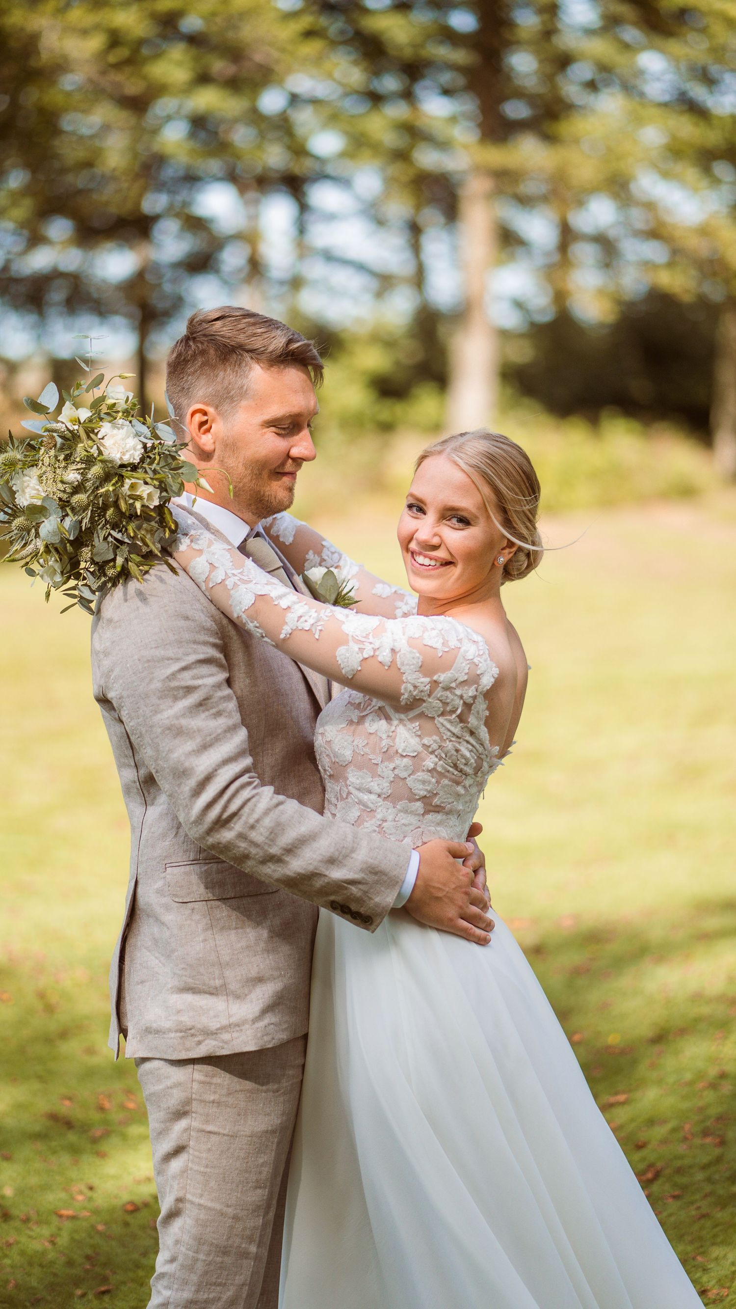 Smiling bride in floral lace gown embraces groom in grey suit while posing in lush green garden setting.
