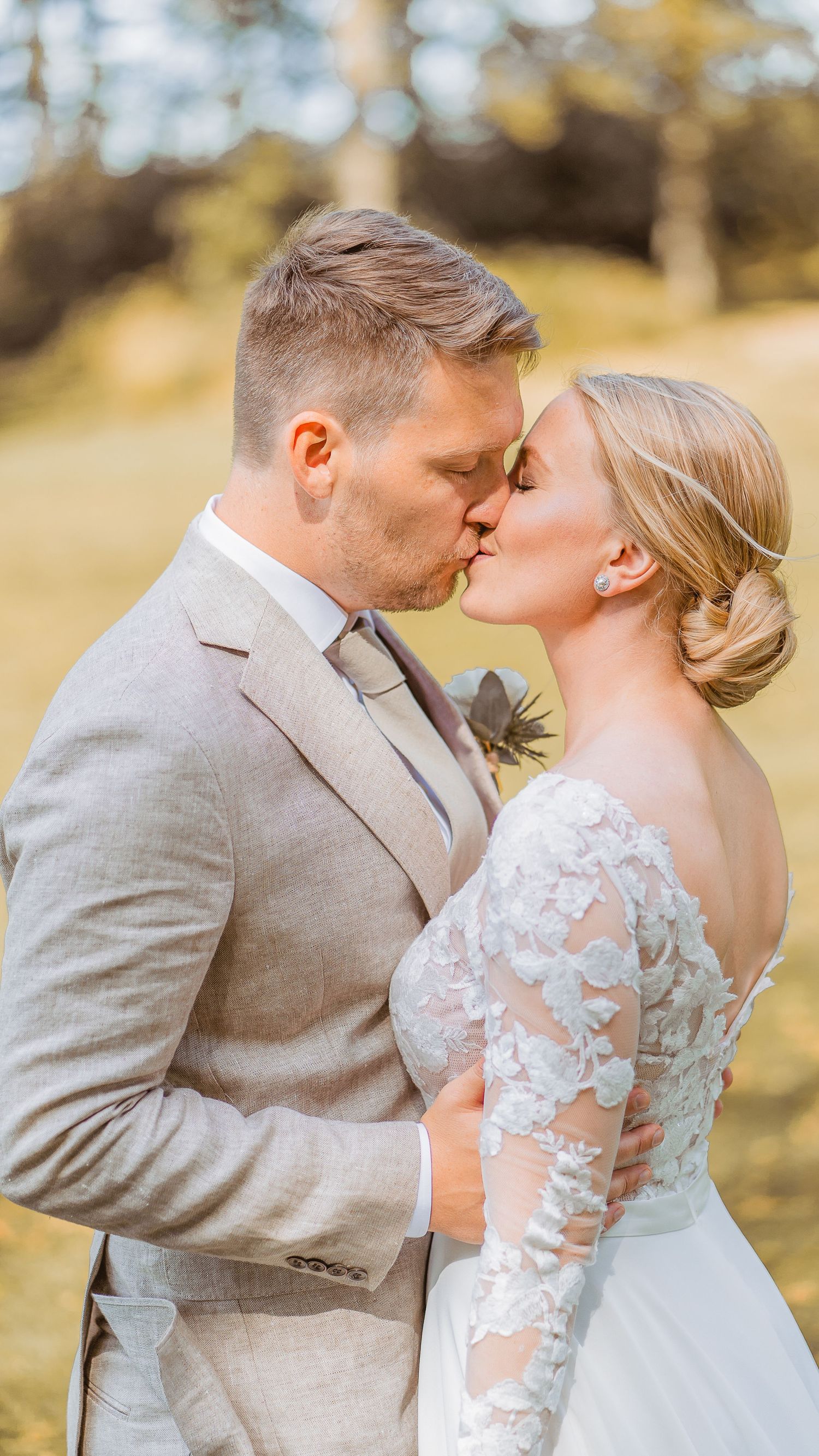Bride and groom share a romantic kiss outdoors in golden light, dressed in lace gown and beige suit.