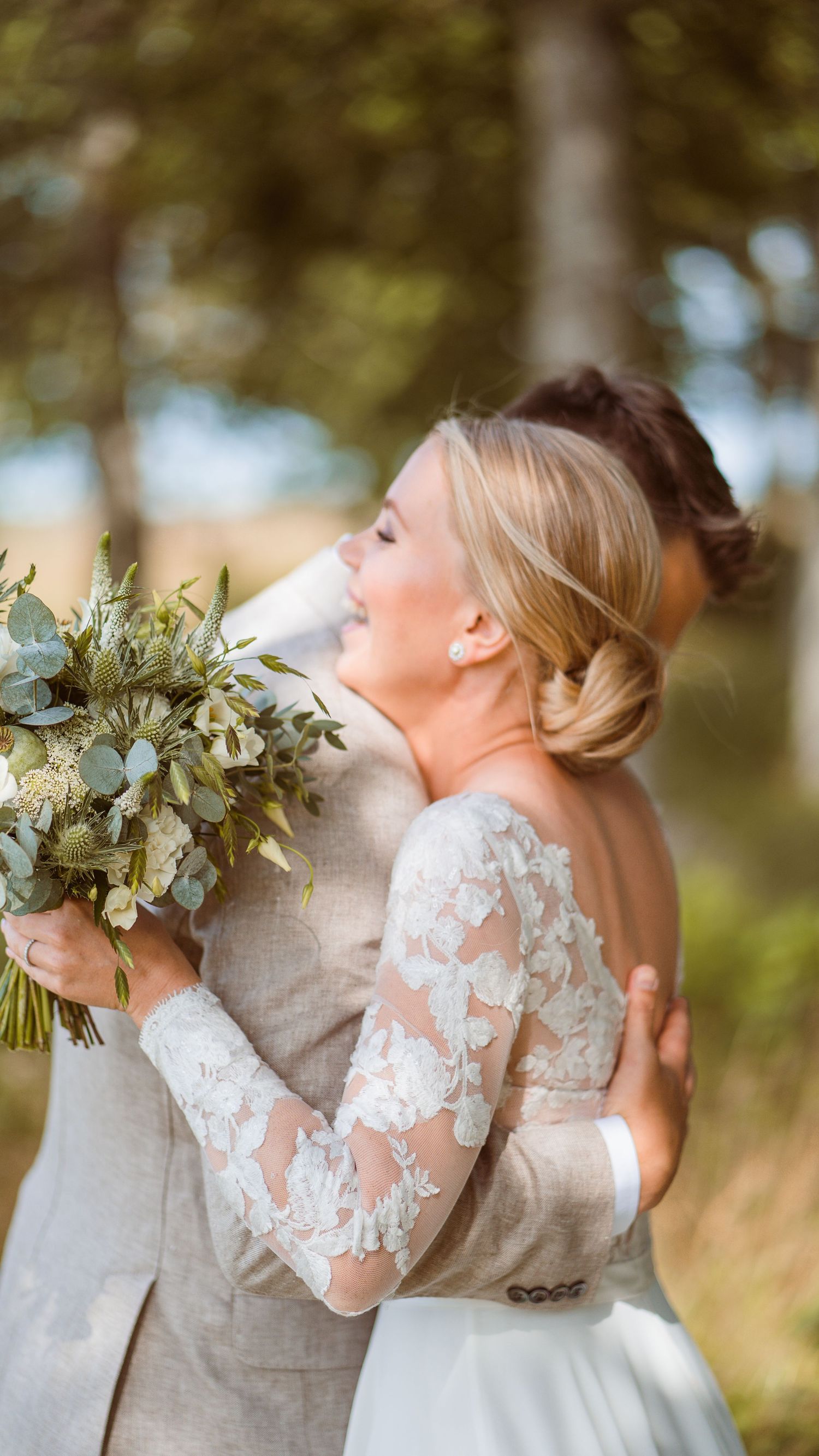 Bride in long-sleeve lace wedding gown embraces groom tenderly while holding white and green floral bouquet.