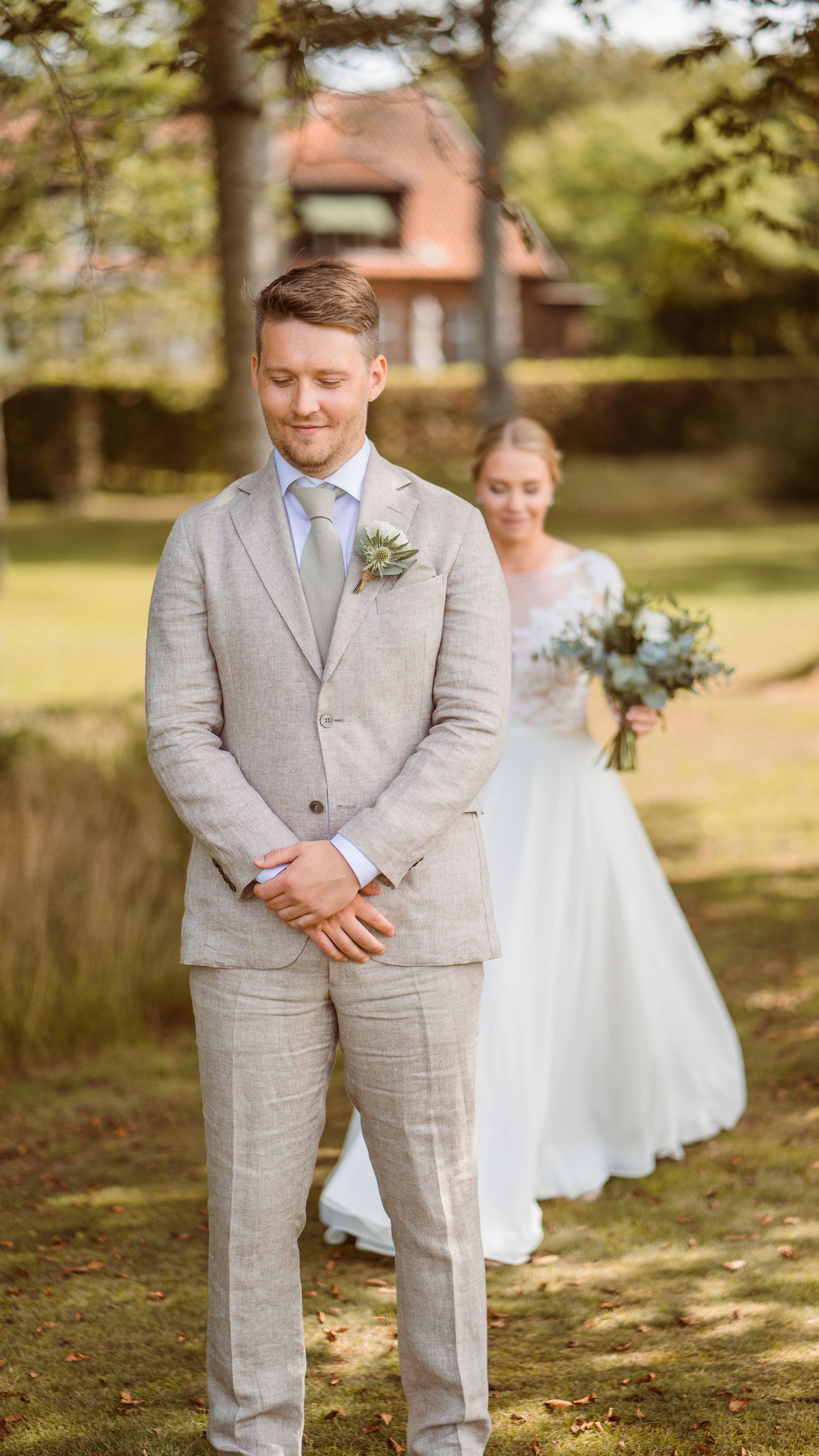 Groom in beige linen suit stands with eyes closed as bride in white lace gown approaches from behind outdoors.
