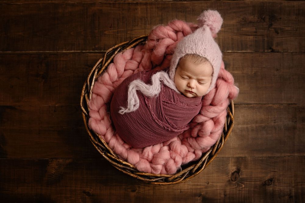 Sleeping newborn wrapped in burgundy fabric nestled in pink bedding inside a wooden basket.