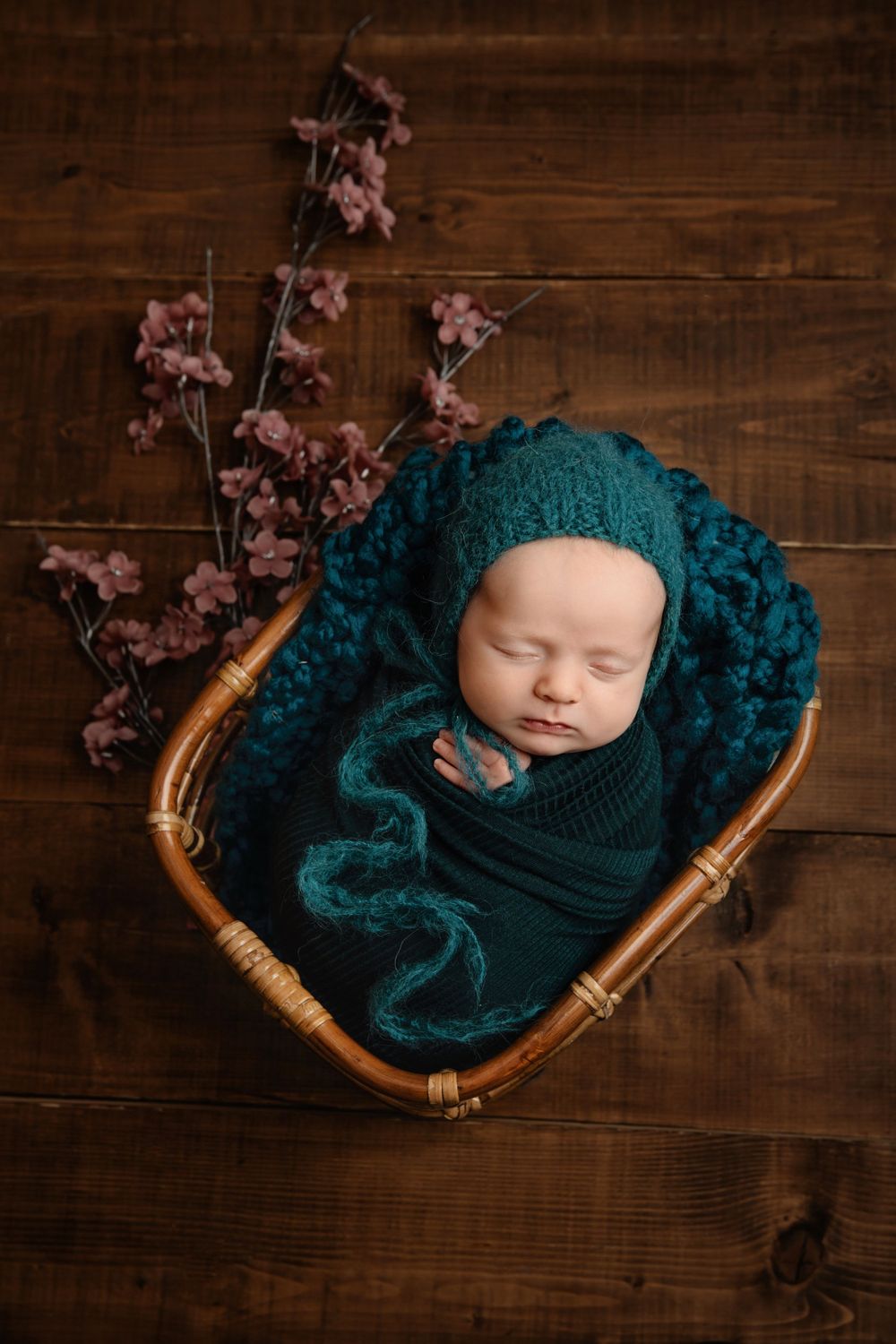 Newborn baby sleeping in teal wrap inside wicker basket with cherry blossoms on wooden background.