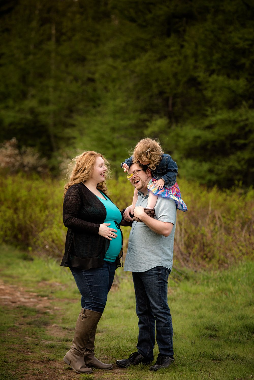 Two expectant parents share a joyful moment outdoors with a toddler in a forested setting.