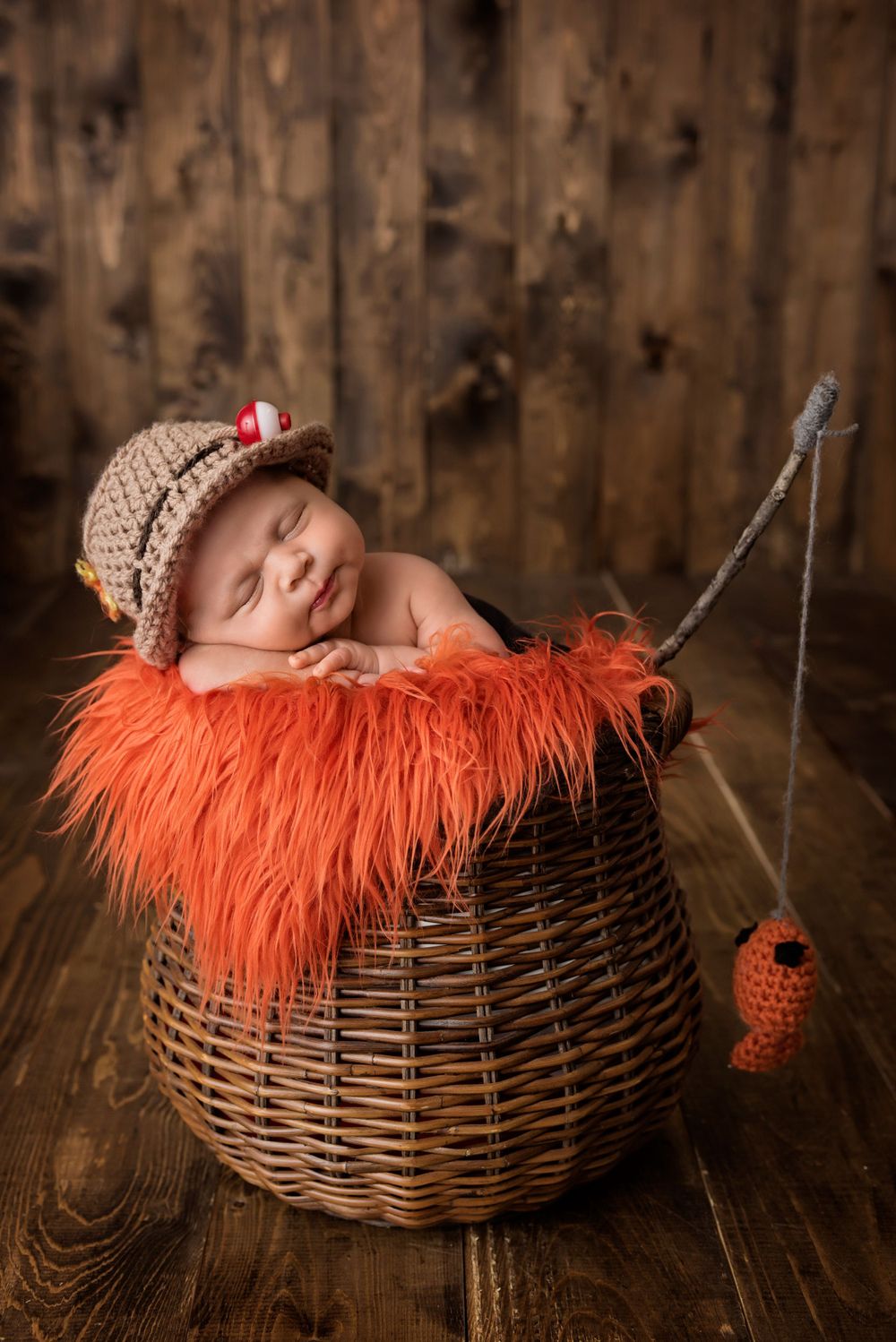 Newborn in knit hat sleeping in fishing basket with orange fur and decorative fishing lure.