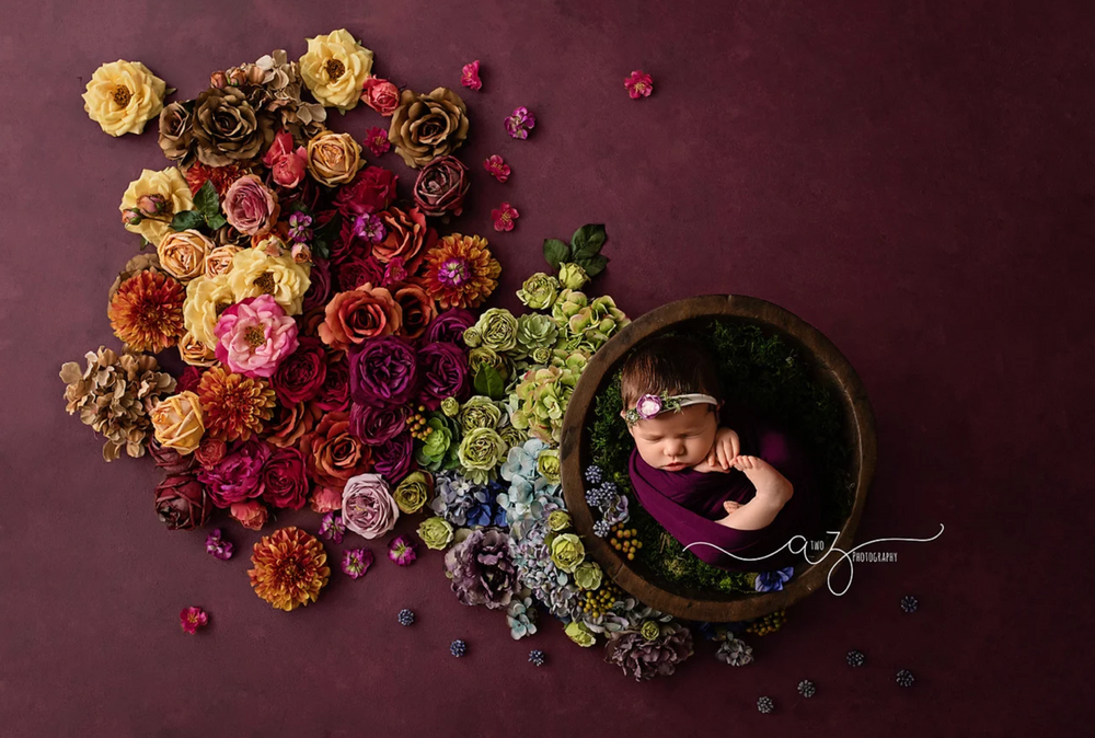 Vibrant floral arrangement with colorful roses and hydrangeas spilling from a round vessel on a burgundy background.