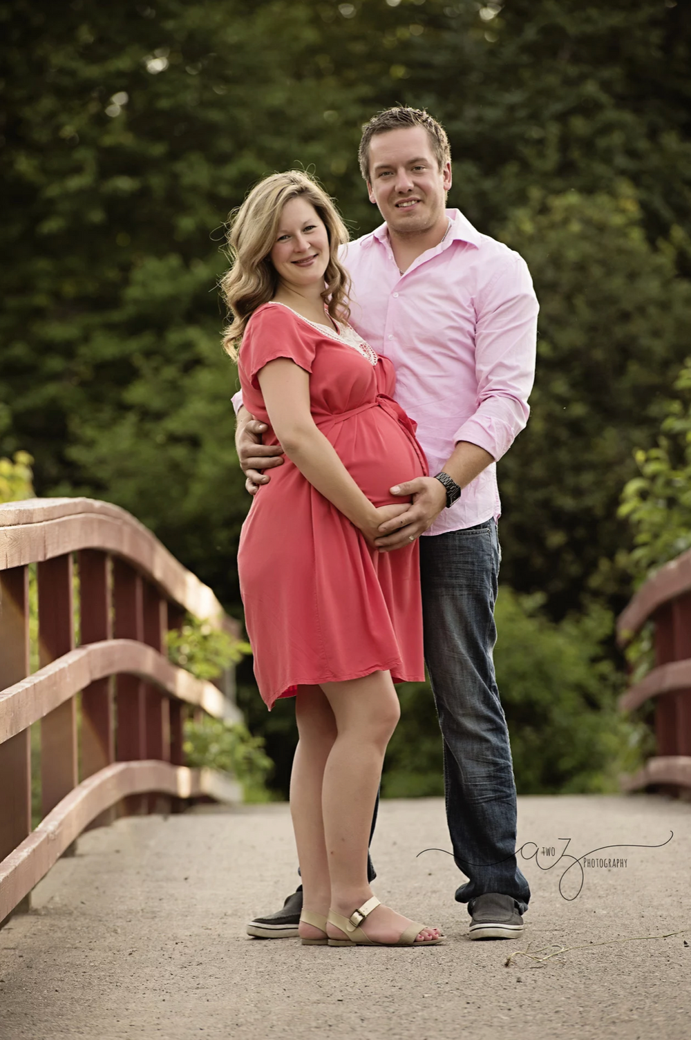 Couple standing on wooden bridge with expecting mother wearing coral dress.