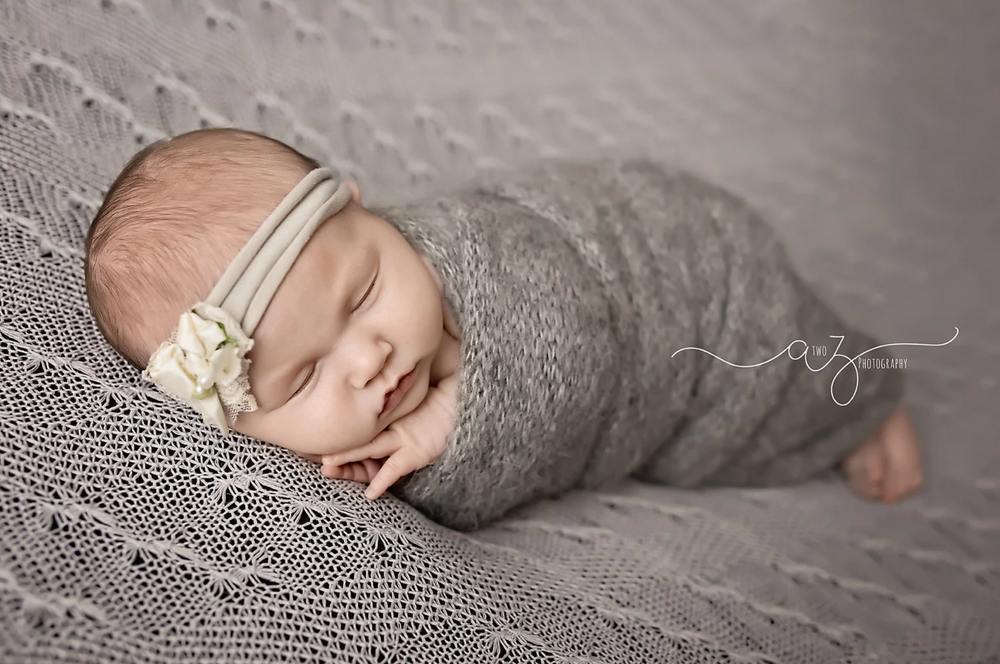 Sleeping newborn wrapped in gray swaddle with lace headband on textured blanket.
