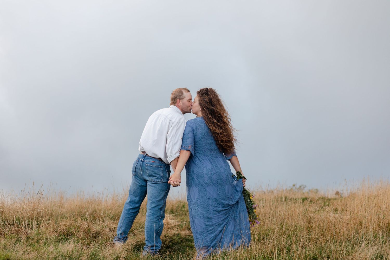Couple shares intimate moment in a grassy meadow with mountains in the background.