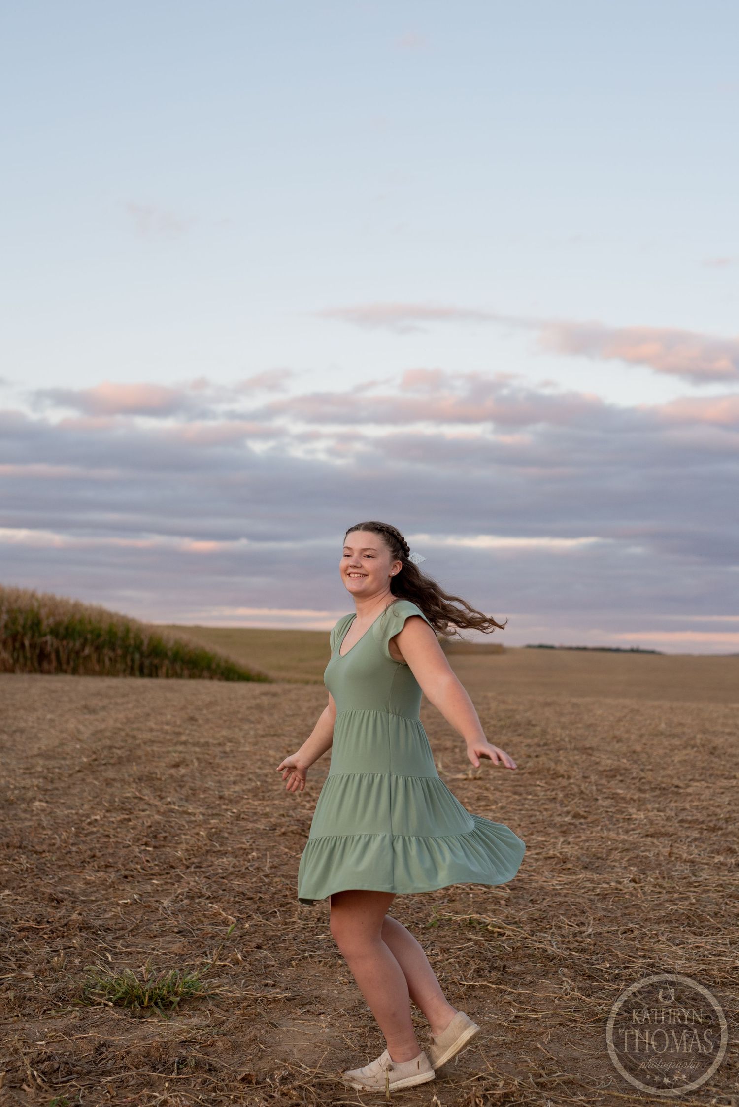 Playful twirling motion captured in a sage dress against sunset clouds and rolling hills.