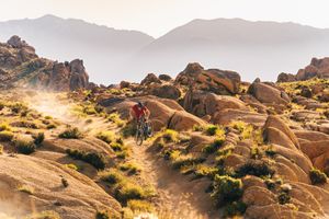 Mountain bikers traverse a winding trail through golden desert rocks and scrub brush under rugged mountains.