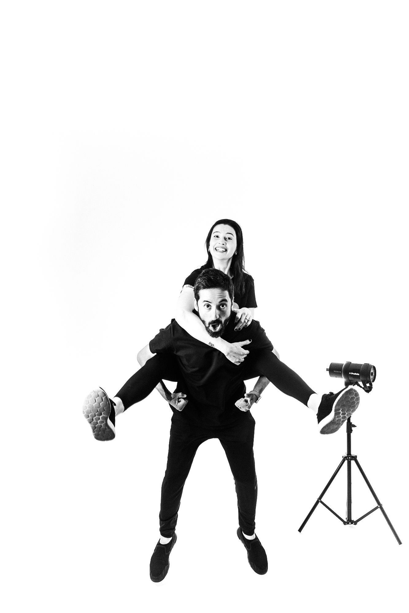 Playful black and white studio portrait of couple in piggyback pose with camera equipment.