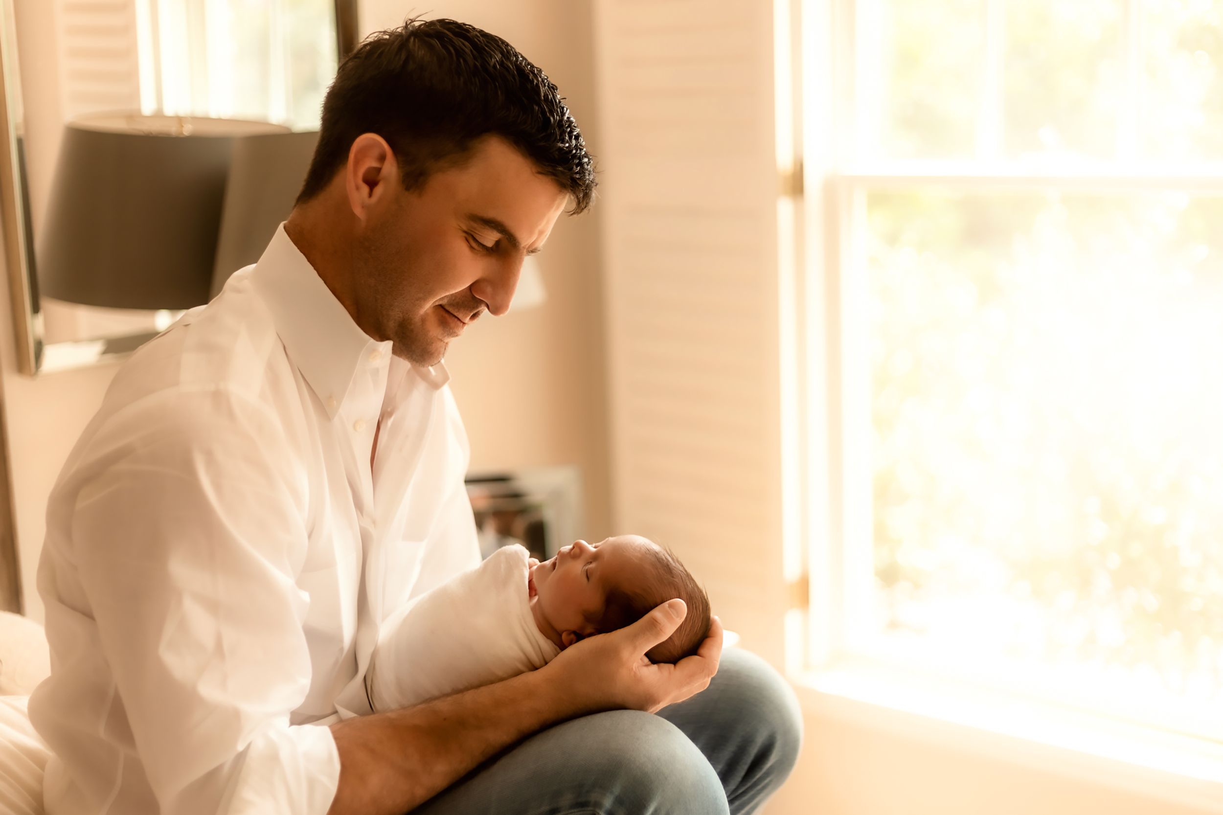 Parent in white shirt tenderly holding sleeping newborn baby by window.