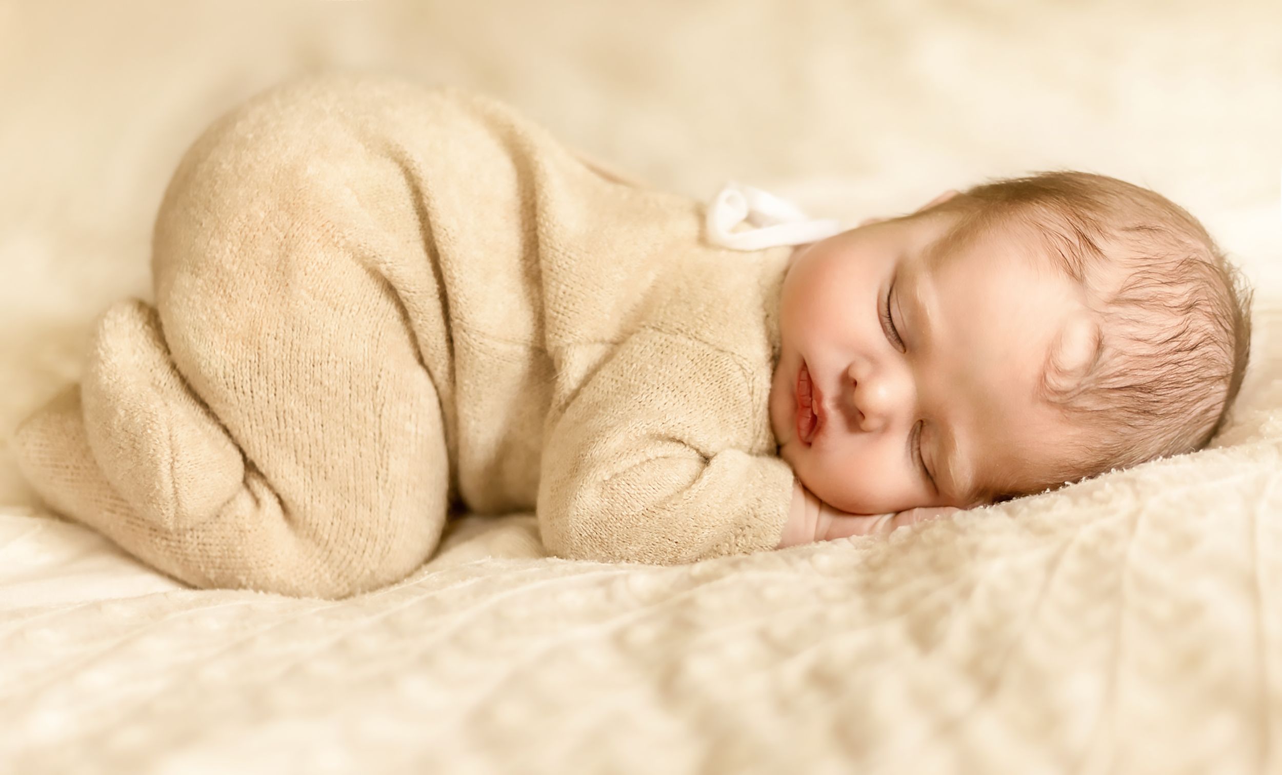 Sleeping newborn baby wrapped in beige knit blanket on cream textured bedding.
