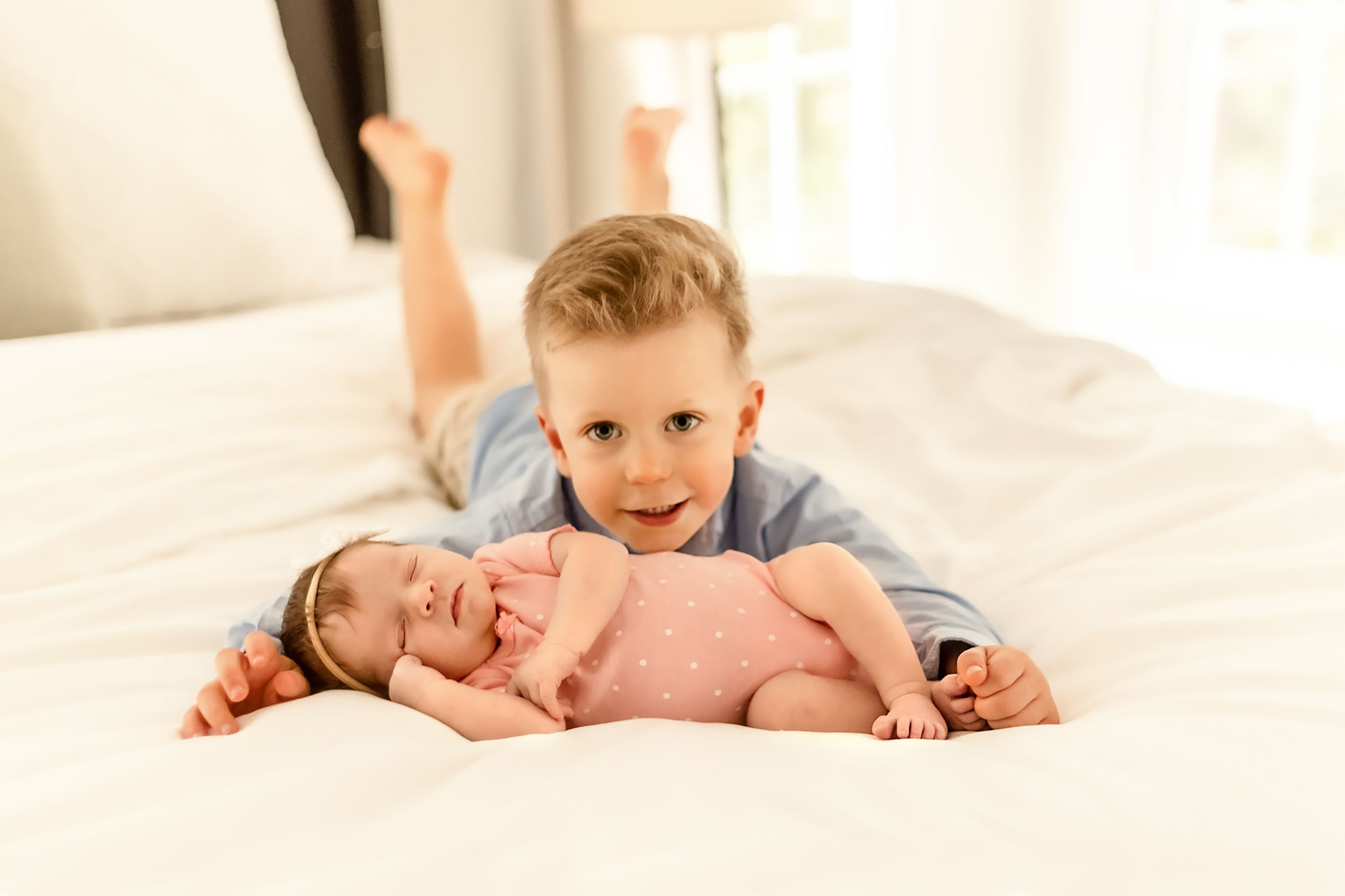 Two children on white bed, older sibling playfully posing with baby.