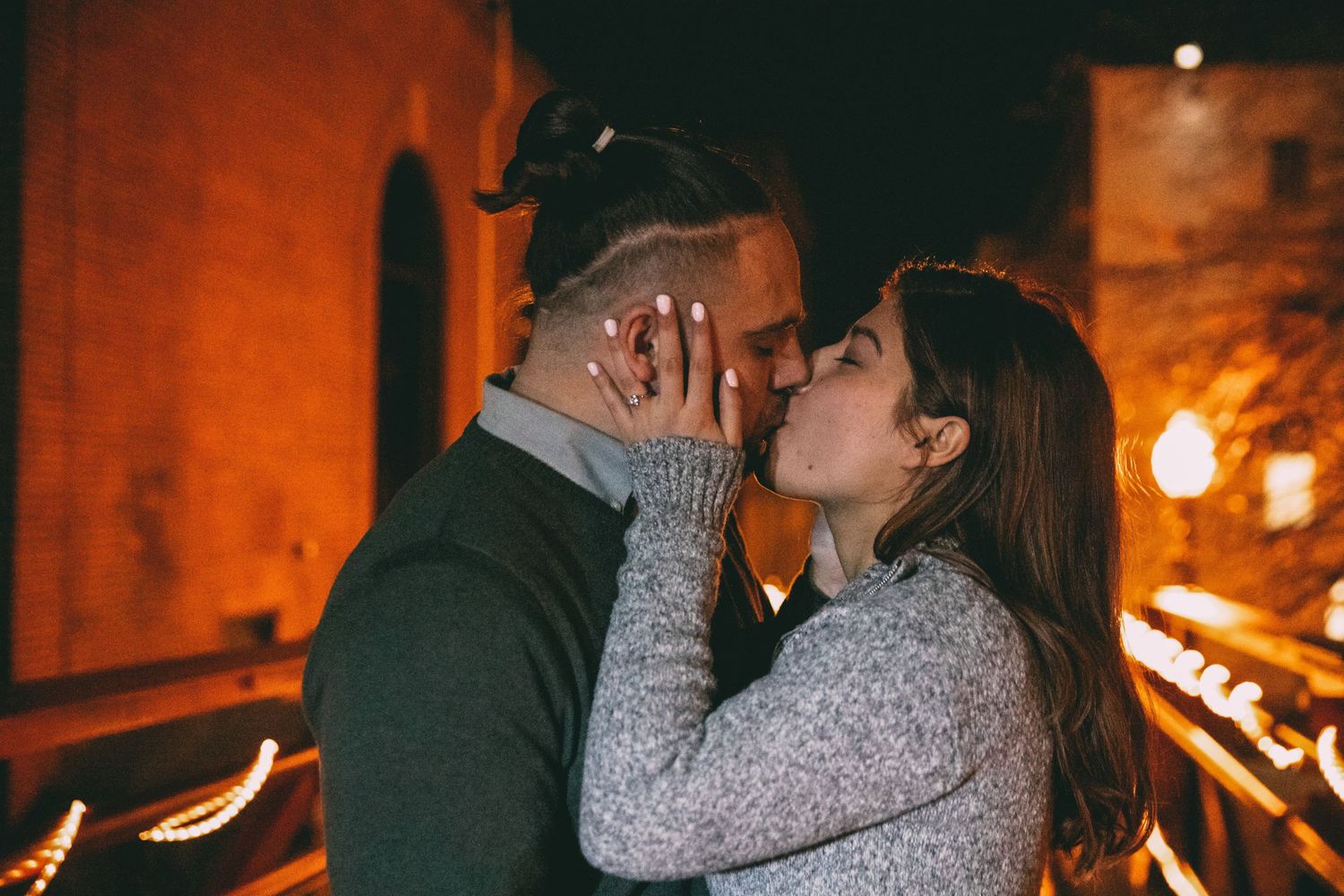 Romantic couple sharing a kiss at night surrounded by warm glowing lights at an outdoor venue.