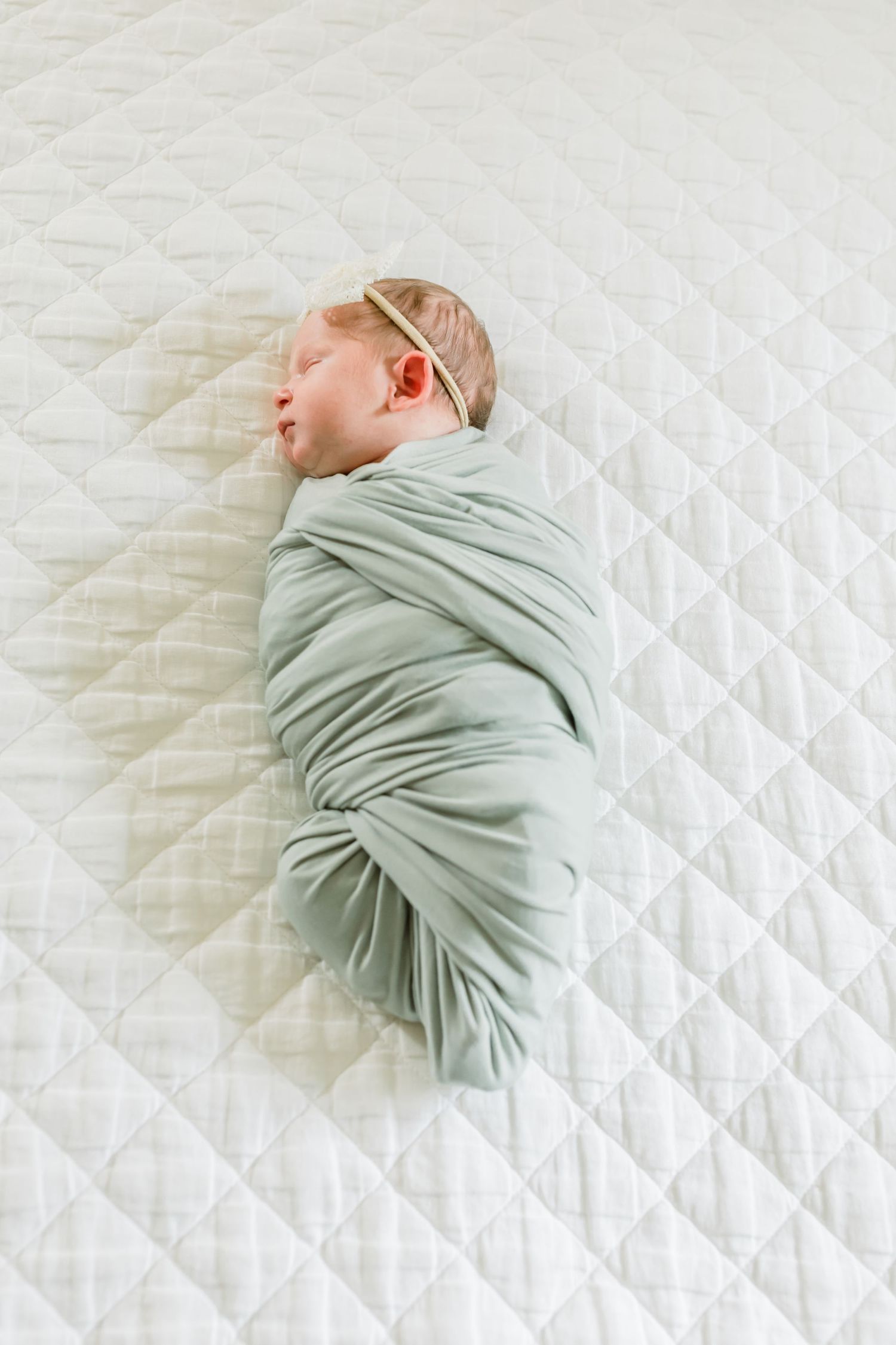 A newborn baby sleeps while swaddled in mint green fabric on a quilted white surface.