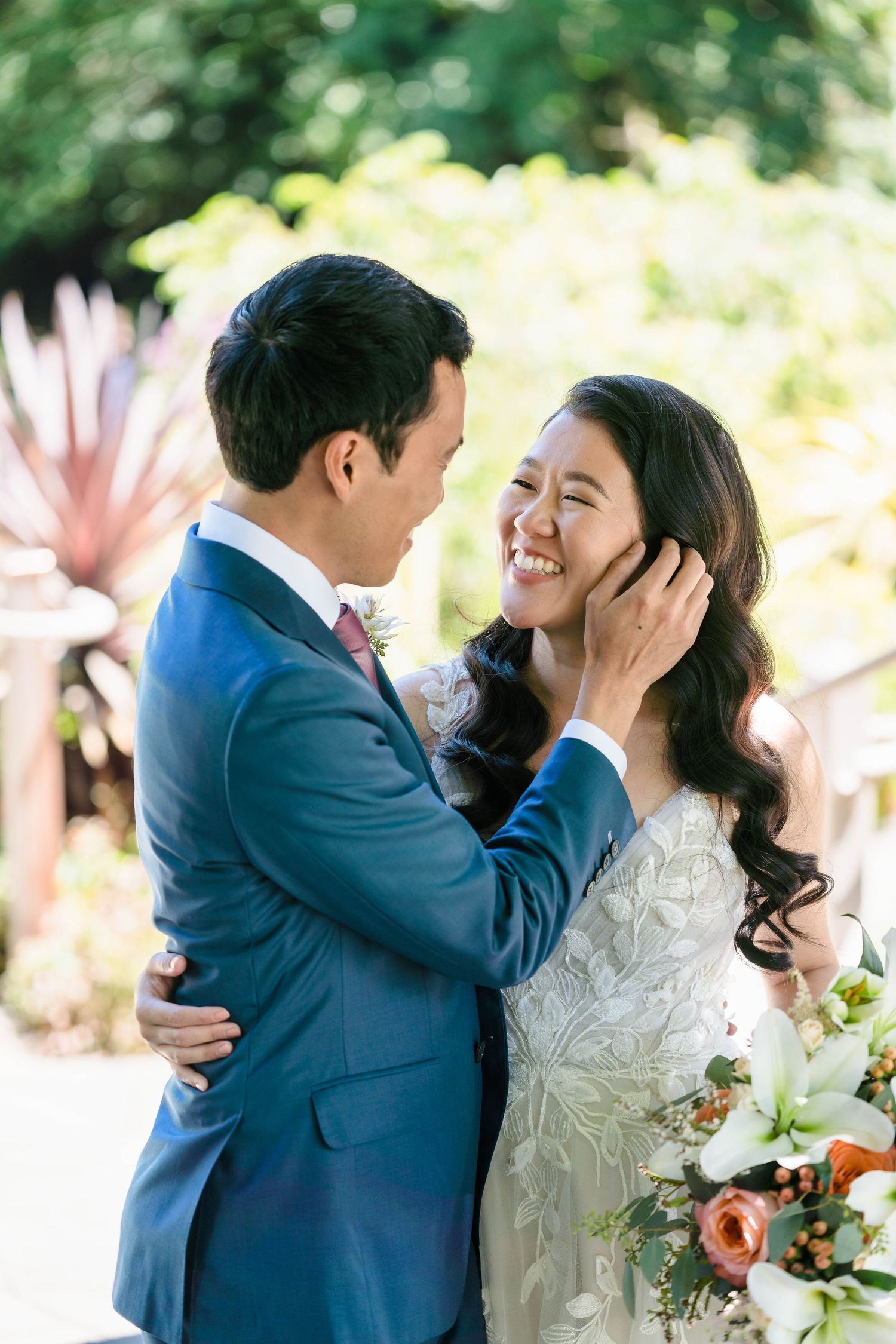 A tender wedding moment captured in a sun-dappled garden with pink flowers.