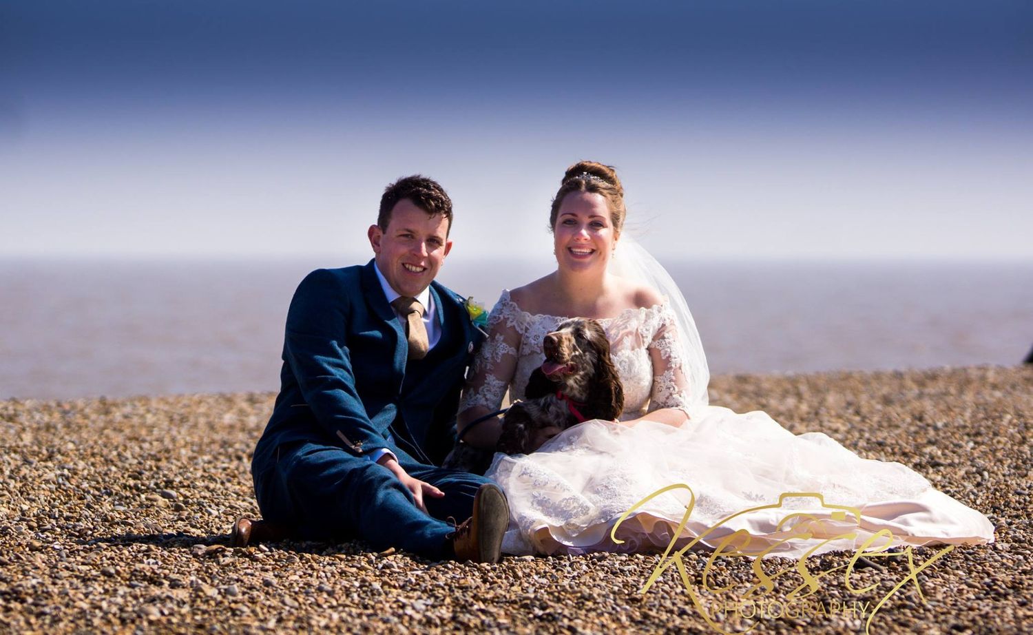 A couple in wedding attire sits on a beach with their dog against a misty backdrop.