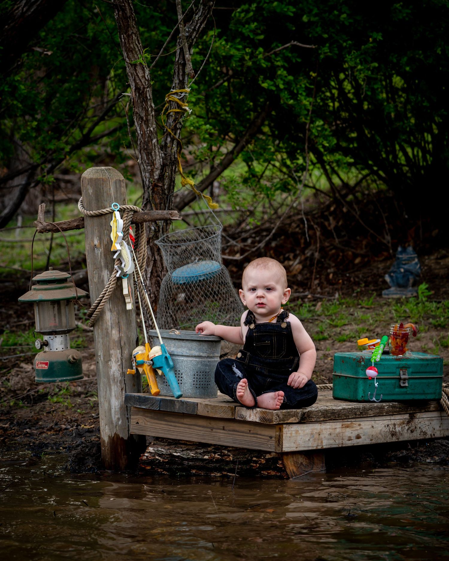 Baby boy in overalls sitting on a small dock with fishing gear near the water.