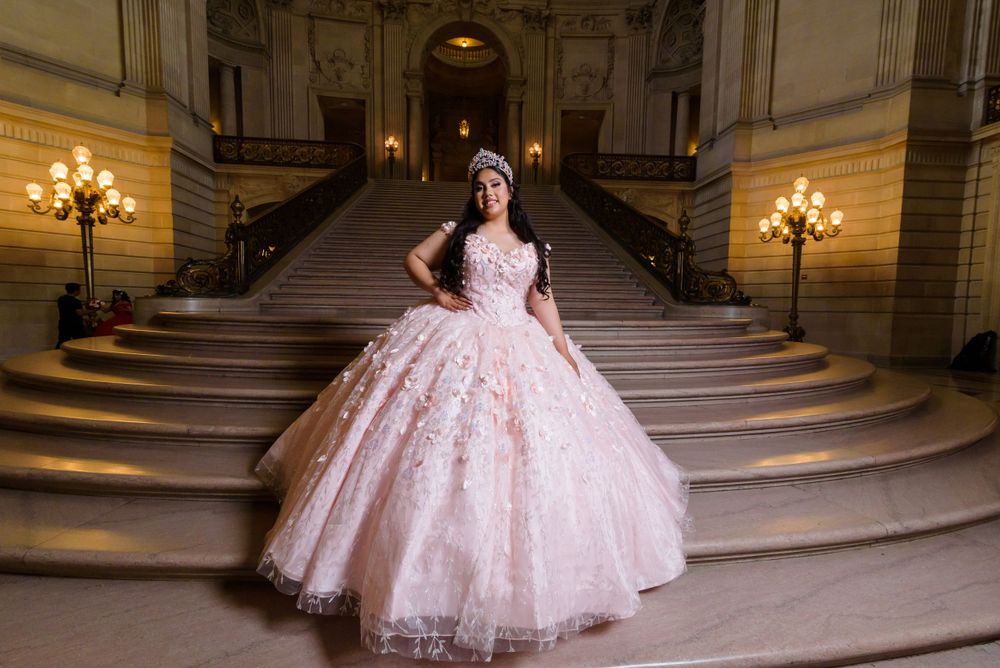 A stunning pink quinceañera ball gown with lace details photographed on an elegant marble staircase with wall sconces.