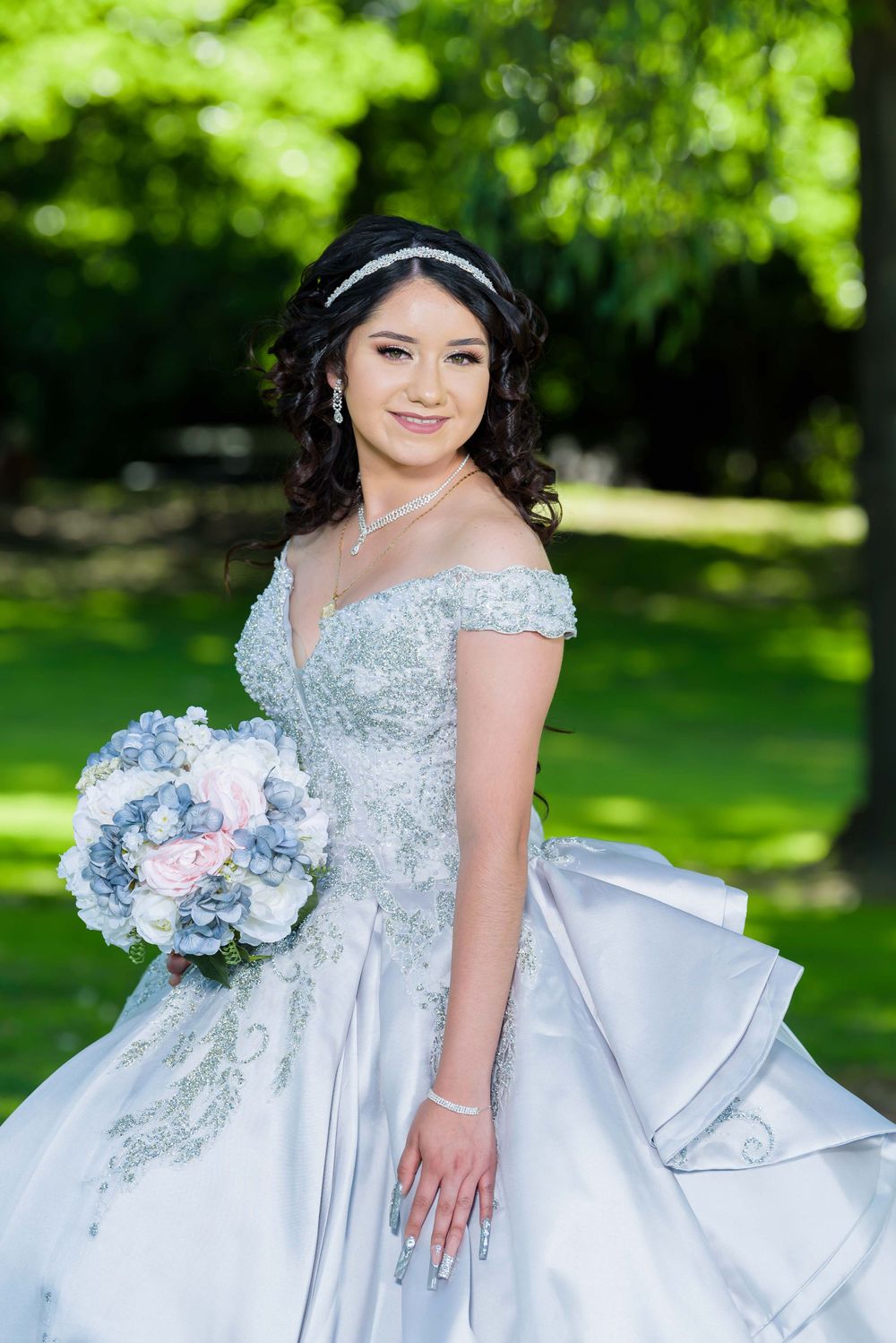 A bride in an elegant off-shoulder silver-white gown holds a blue and pink bouquet in a garden setting.